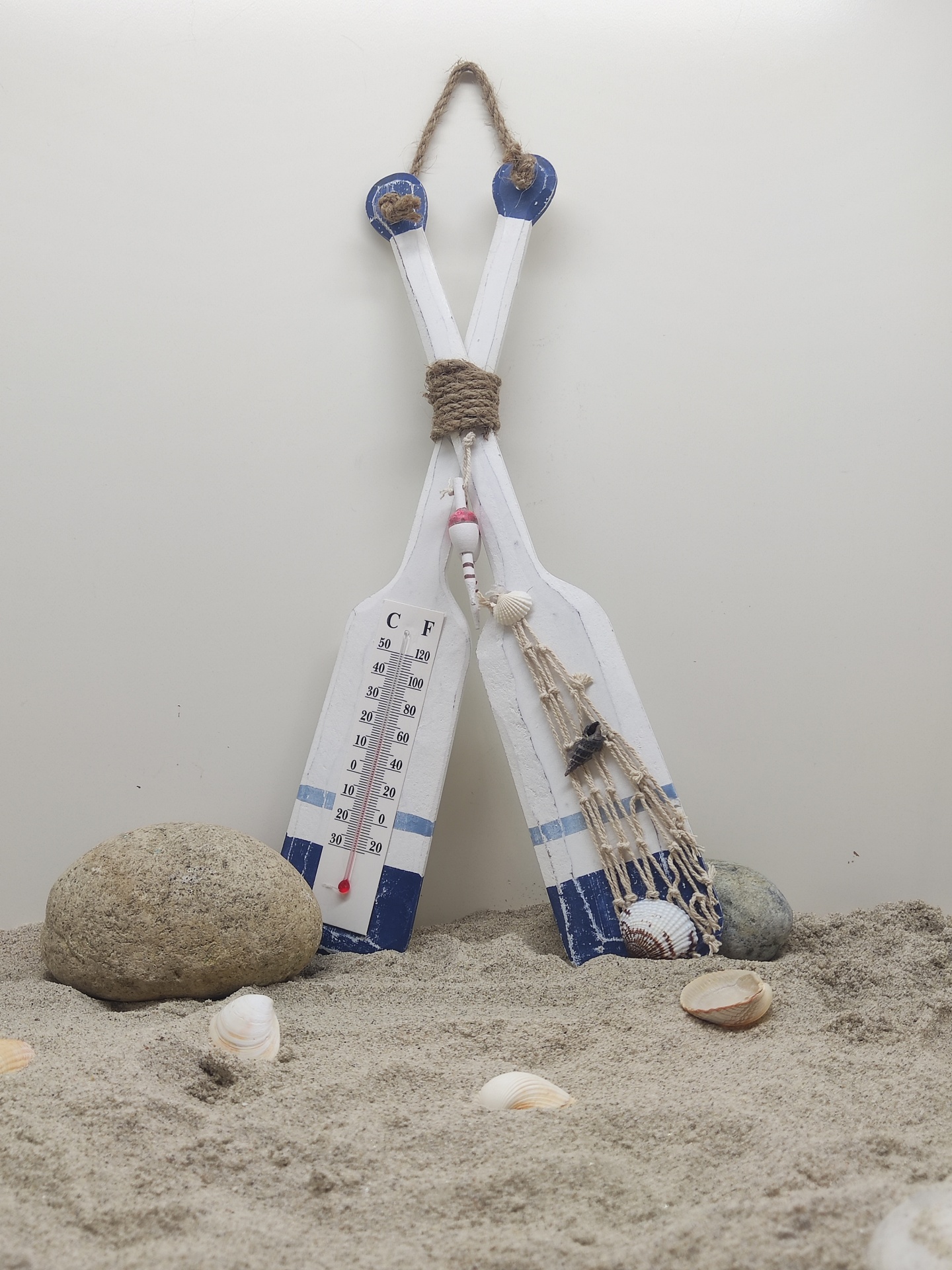 Dois remos decorativos brancos e azuis com termómetro e rede com conchas em ambiente de areia com pedras
