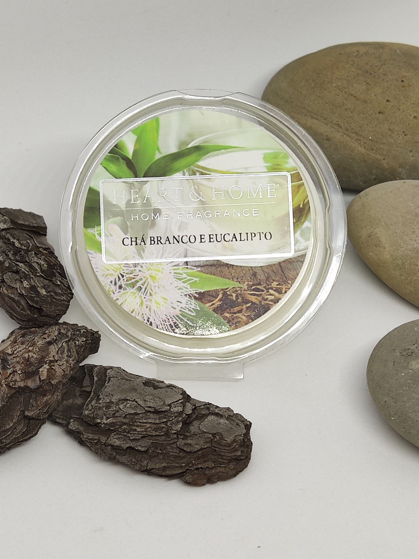 Vela perfumada Chá Branco e Eucalipto em embalagem transparente com etiqueta decorativa, rodeada por casca de árvore e pedras