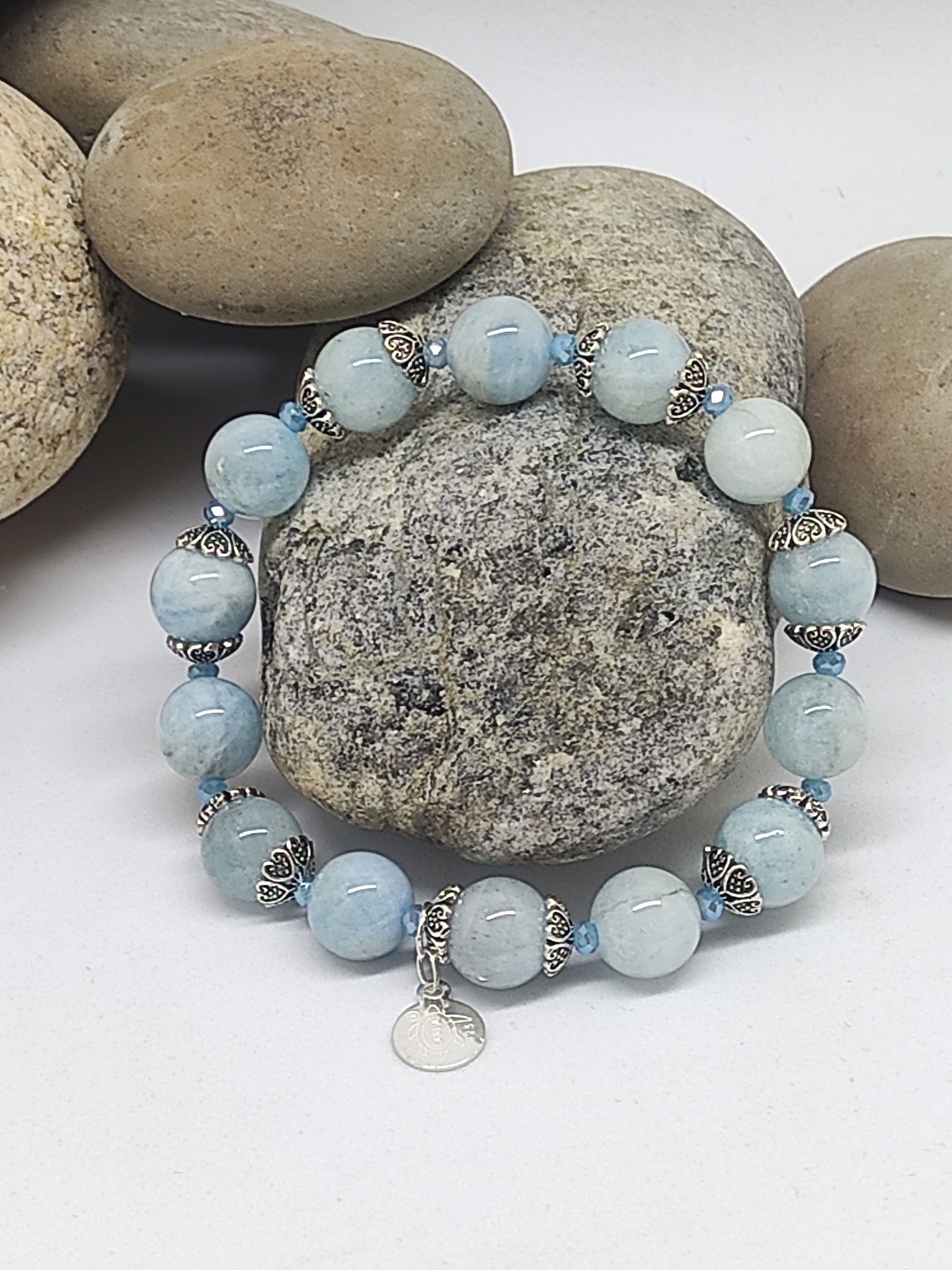 Pulseira azul clara com espaçadores metálicos prateados e pendente, sobre pedras naturais.