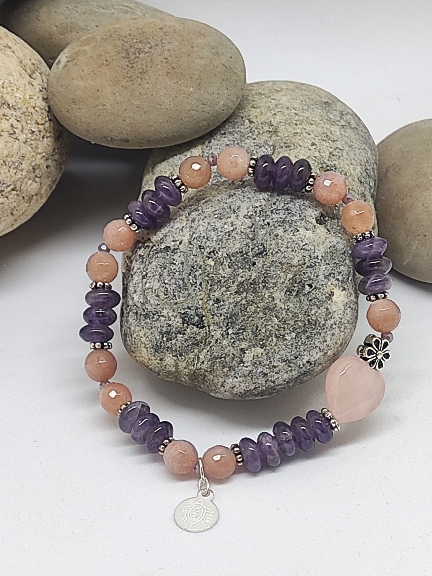 Pulseira de pedras em tons castanho-avermelhado e roxo com pendente metálico, sobre pedras lisas cinzentas