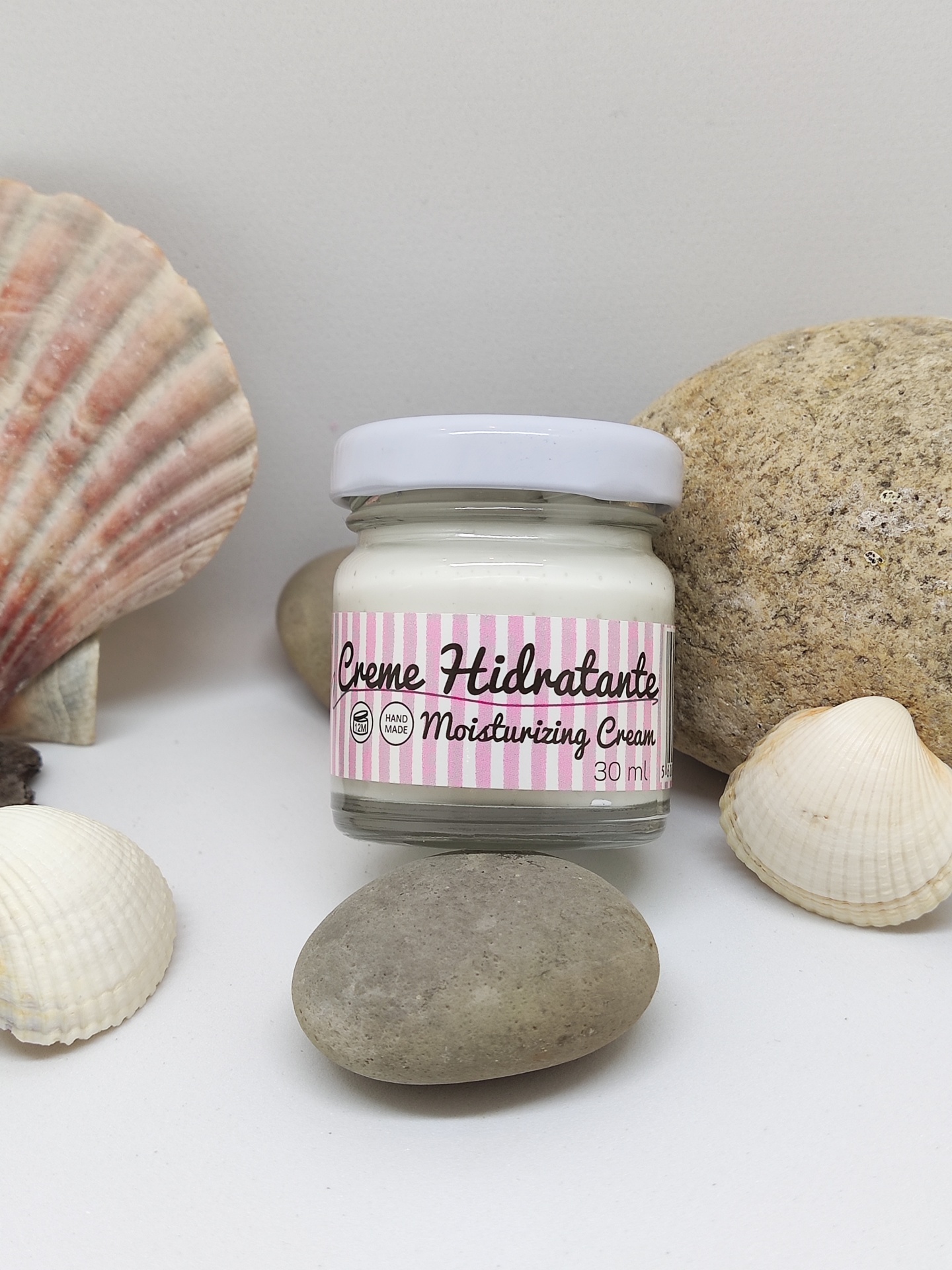 Frasco de creme hidratante em vidro com rótulo rosa e branco, colocado entre pedras e conchas