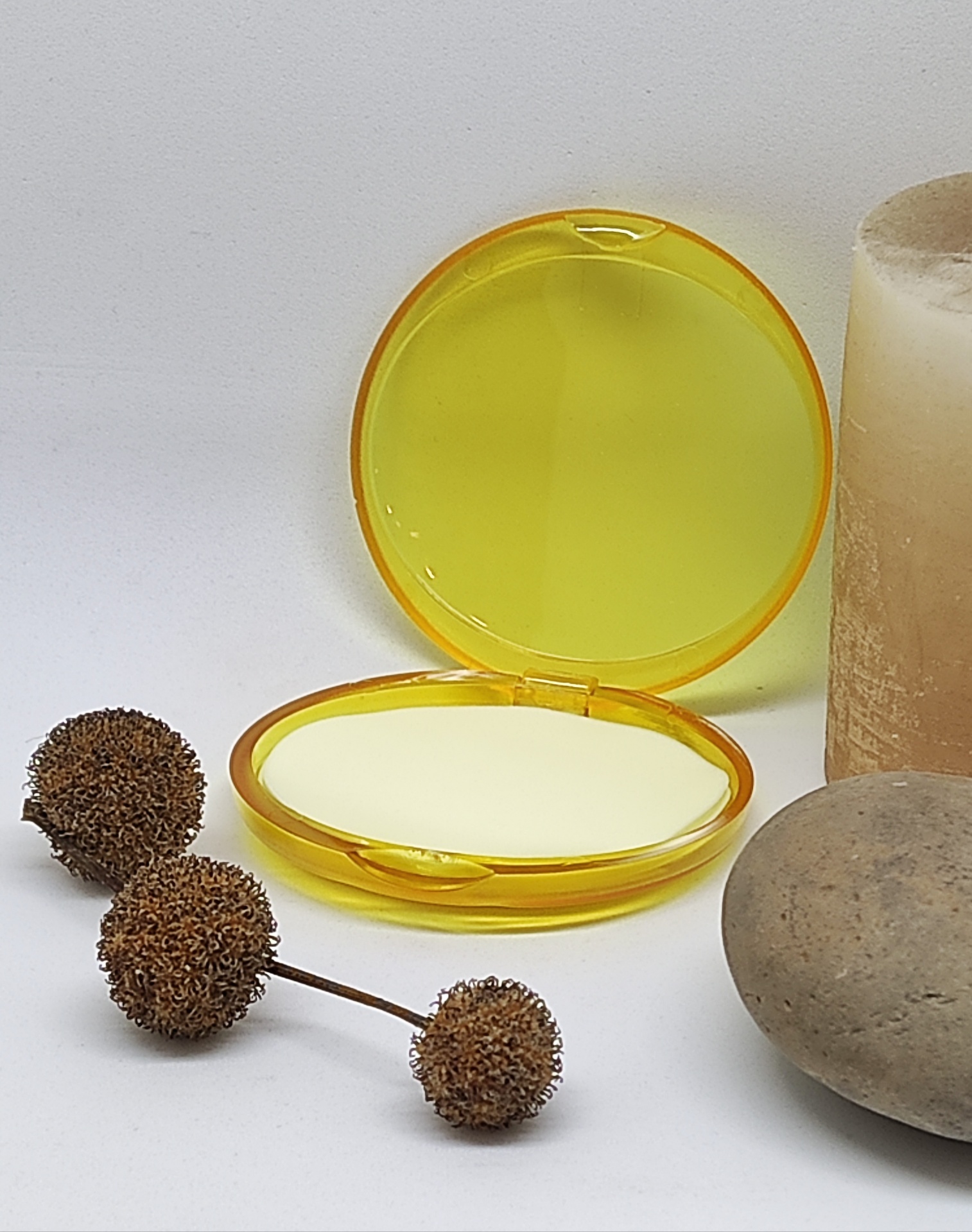 Estojo redondo amarelo translúcido aberto com espelho e base branca ao lado de pedras e flores secas