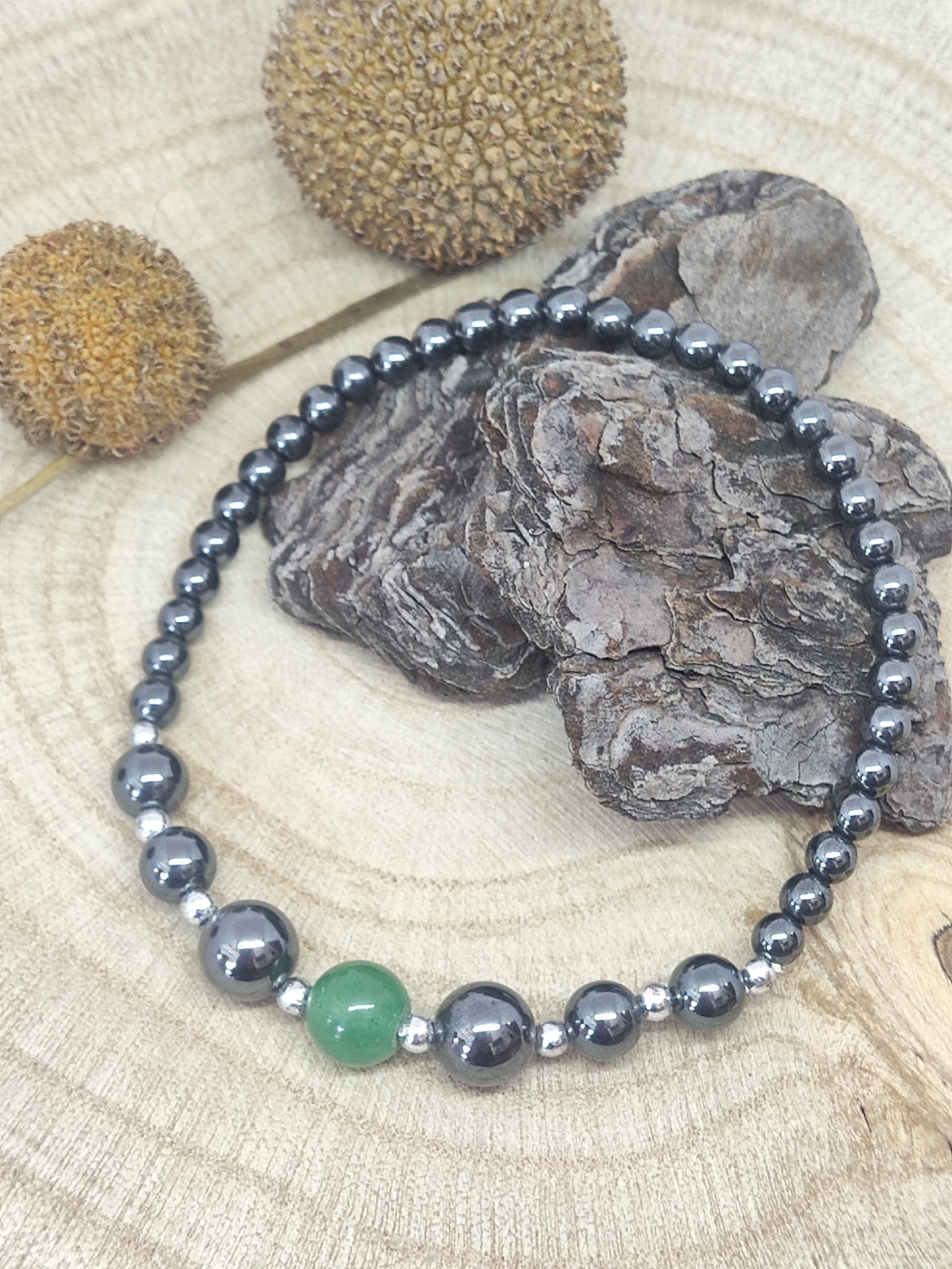 Pulseira de contas metálicas prateadas com conta verde central sobre madeira, casca e elementos naturais secos