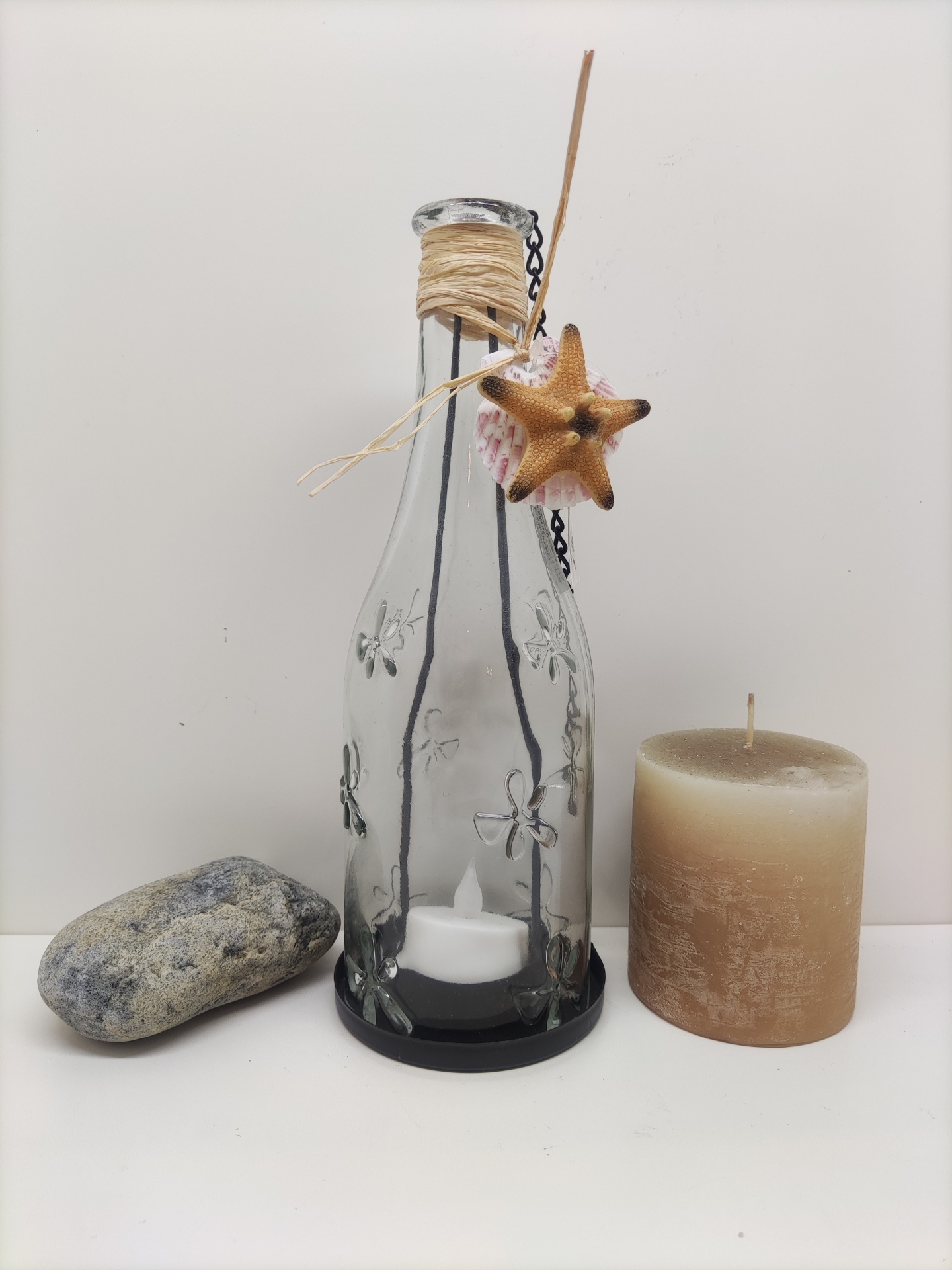 Garrafa de vidro decorada com flor em relevo e corda, estrela-do-mar, concha, pedra cinzenta e vela bege sobre superfície branca