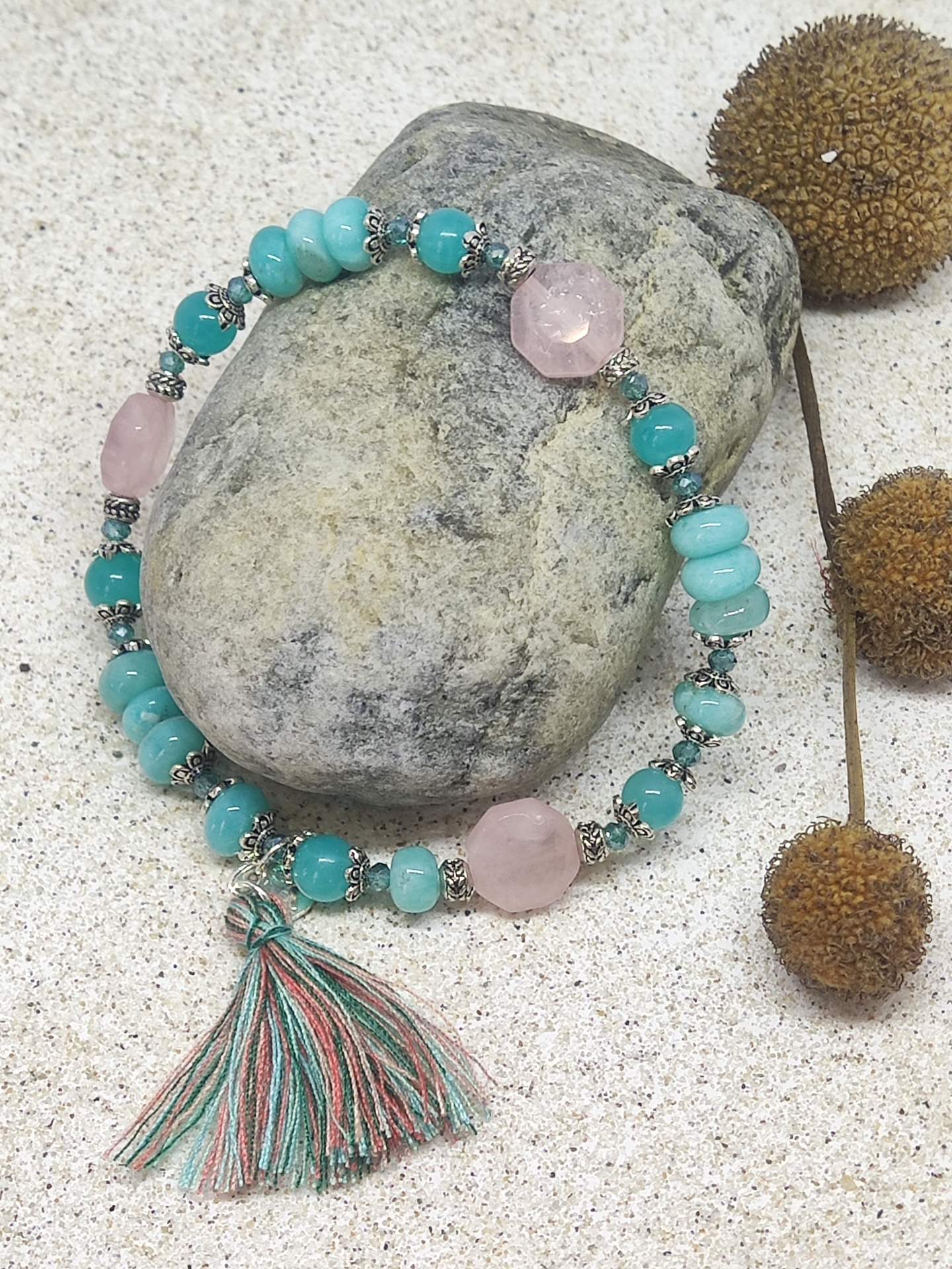 Pulseira de contas azuis e rosas com pendente de fio sobre pedra cinzenta em fundo de areia clara