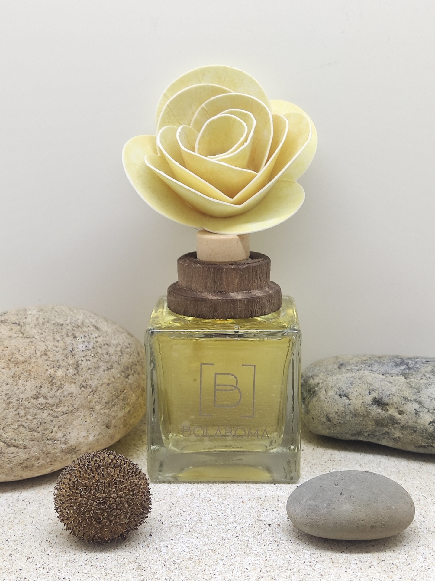 Frasco de vidro quadrado com flor artificial na tampa, rodeado por pedras