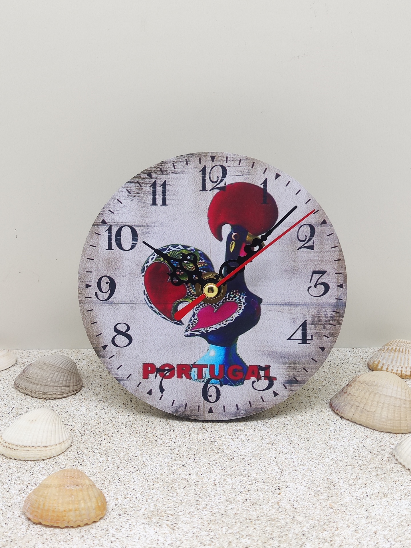 Relógio redondo com imagem de um galo e texto portugal, sobre areia com conchas