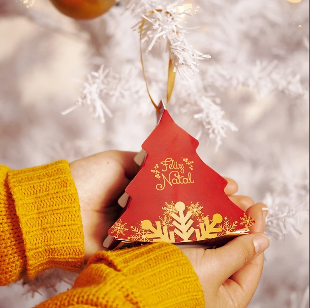 Ornamento de Natal em forma de árvore vermelha com texto 'Feliz Natal' segurado por duas mãos com camisolas amarelas