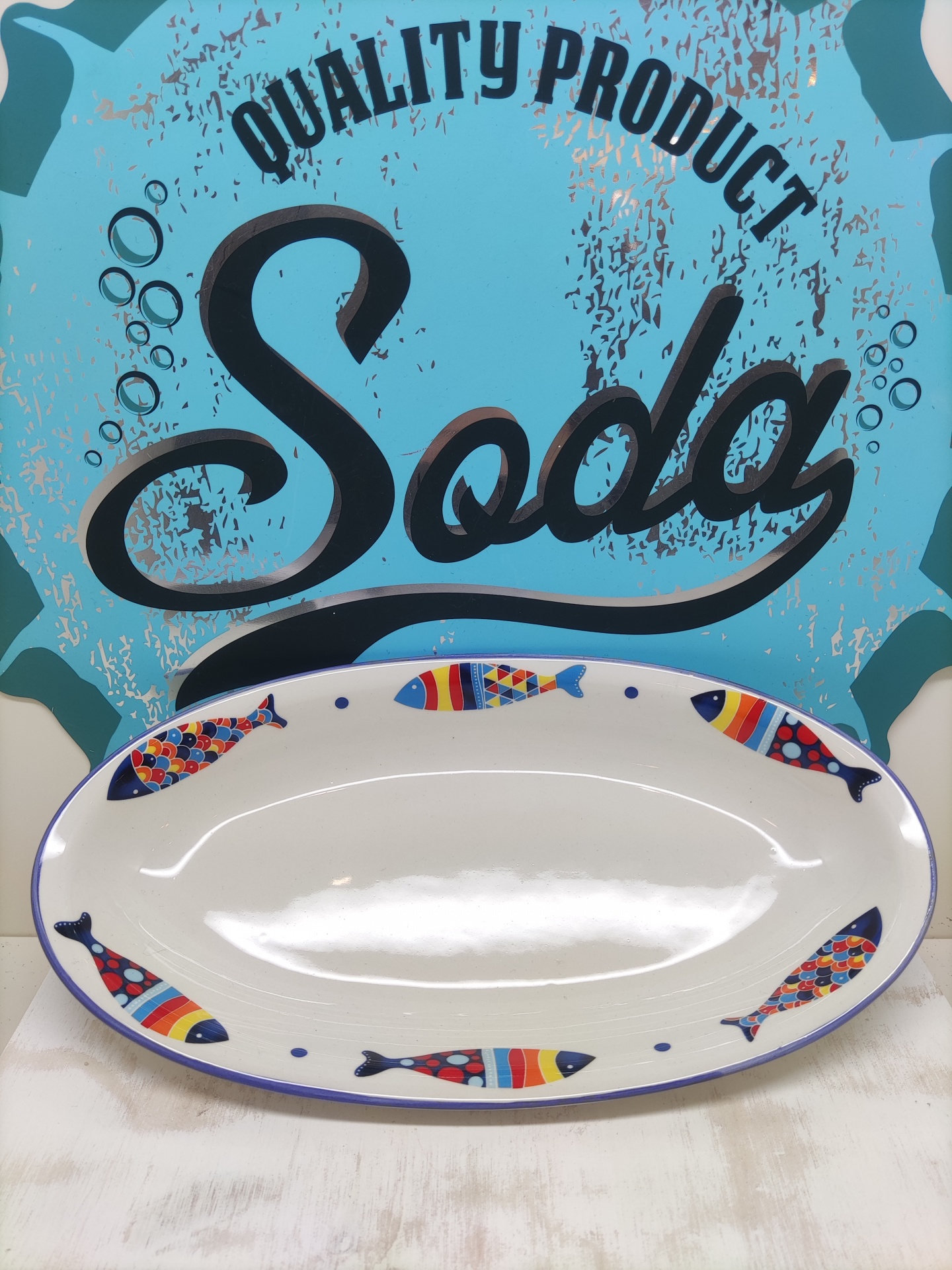 Prato oval branco com peixes coloridos e texto 'QUALITY PRODUCT Soda' no fundo azul