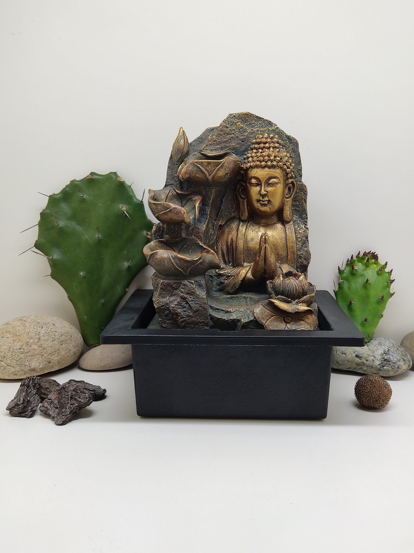 Fonte decorativa com estátua de Buda em tom dourado e base preta junto a cactos e pedras