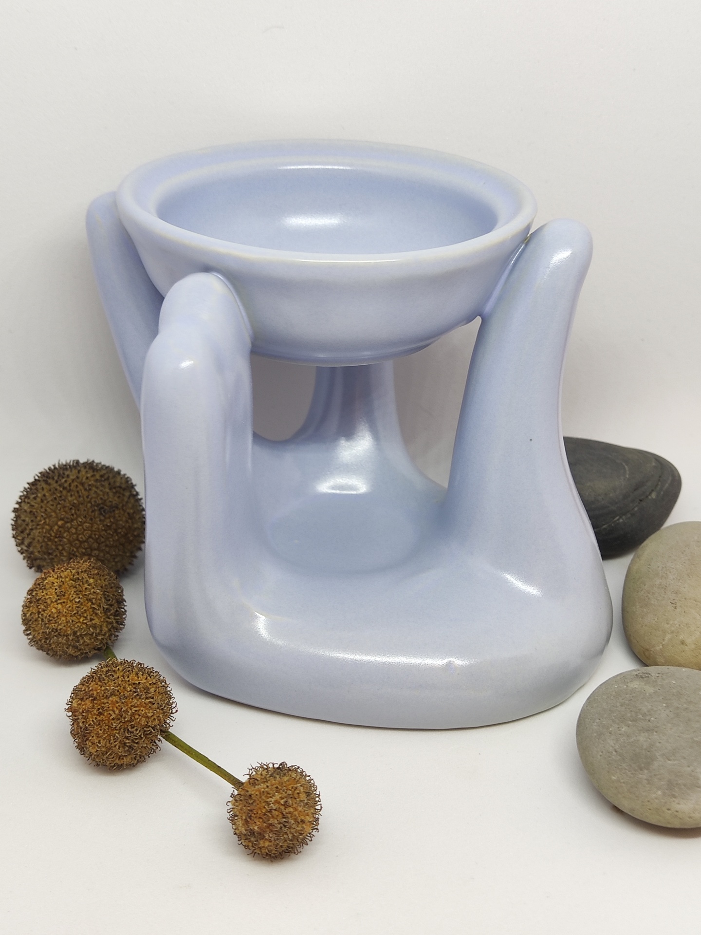 Suporte decorativo azul em cerâmica com formato de mãos e taça, rodeado por pedras e sementes secas.