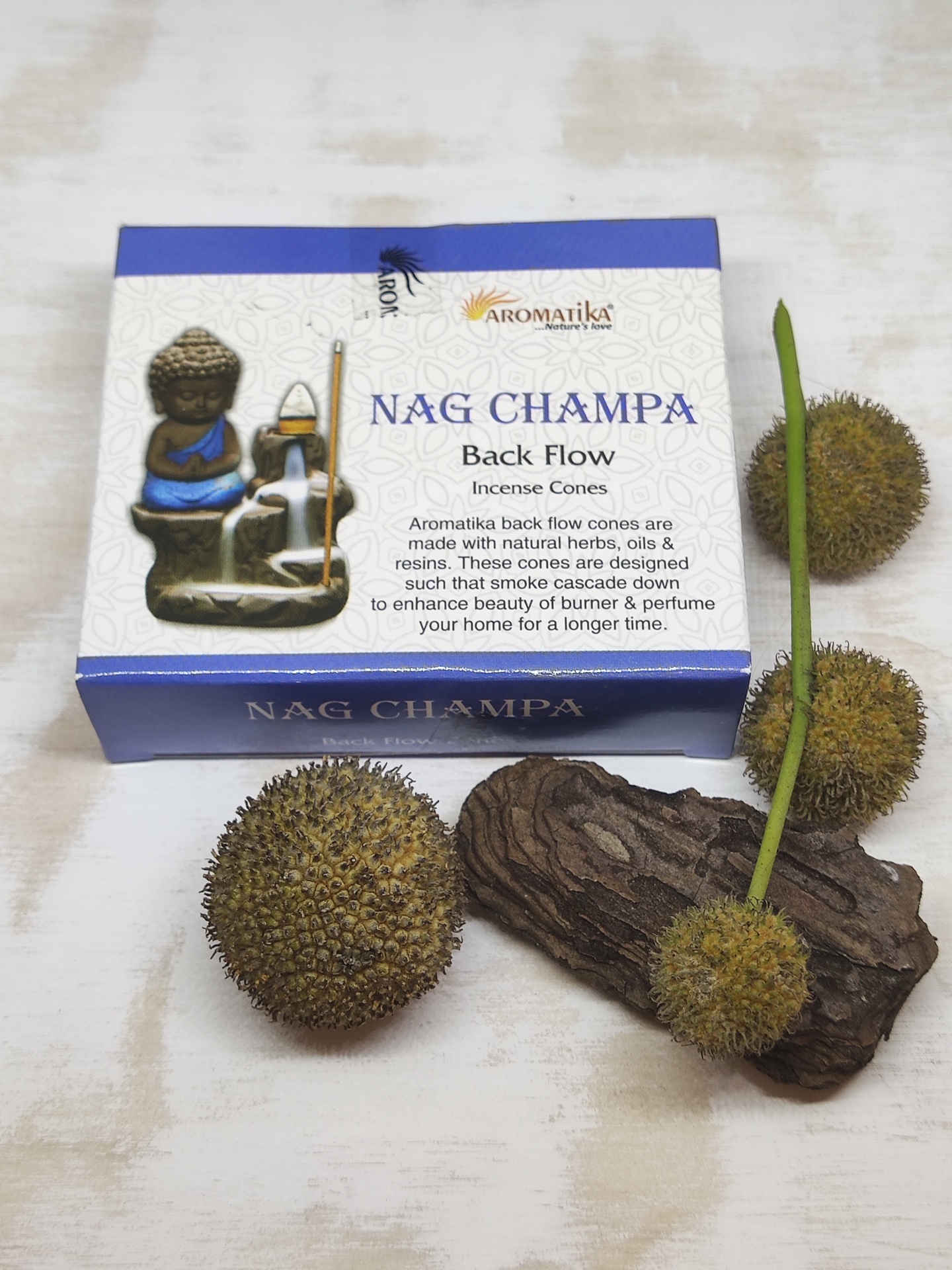 Caixa de incensos Nag Champa Aromatika com estátua de Buda e frutas espinhosas ao lado sobre superfície branca.