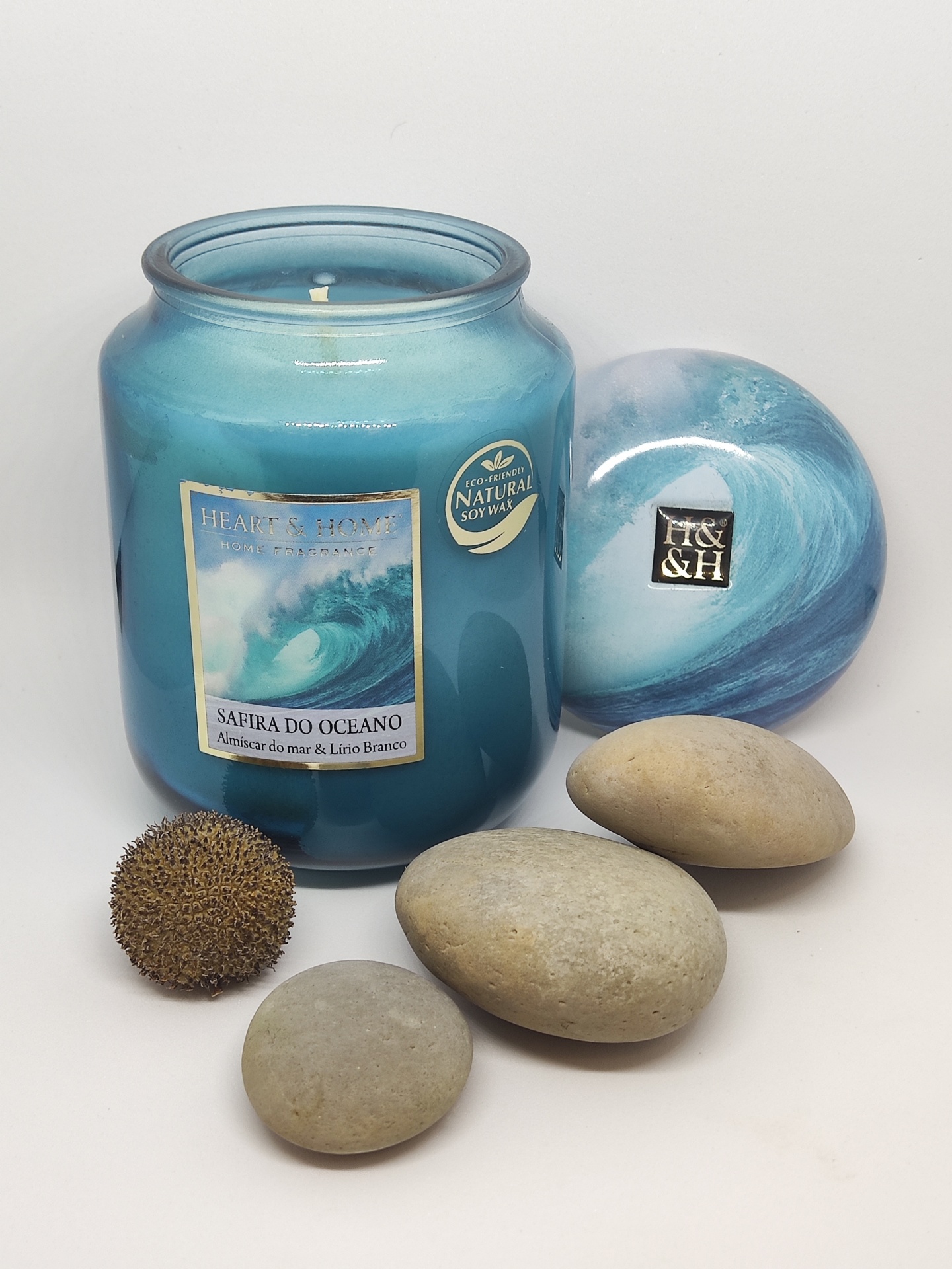 Vela azul em frasco de vidro com tampa azul decorativa e pedras lisas e semente marrom ao lado