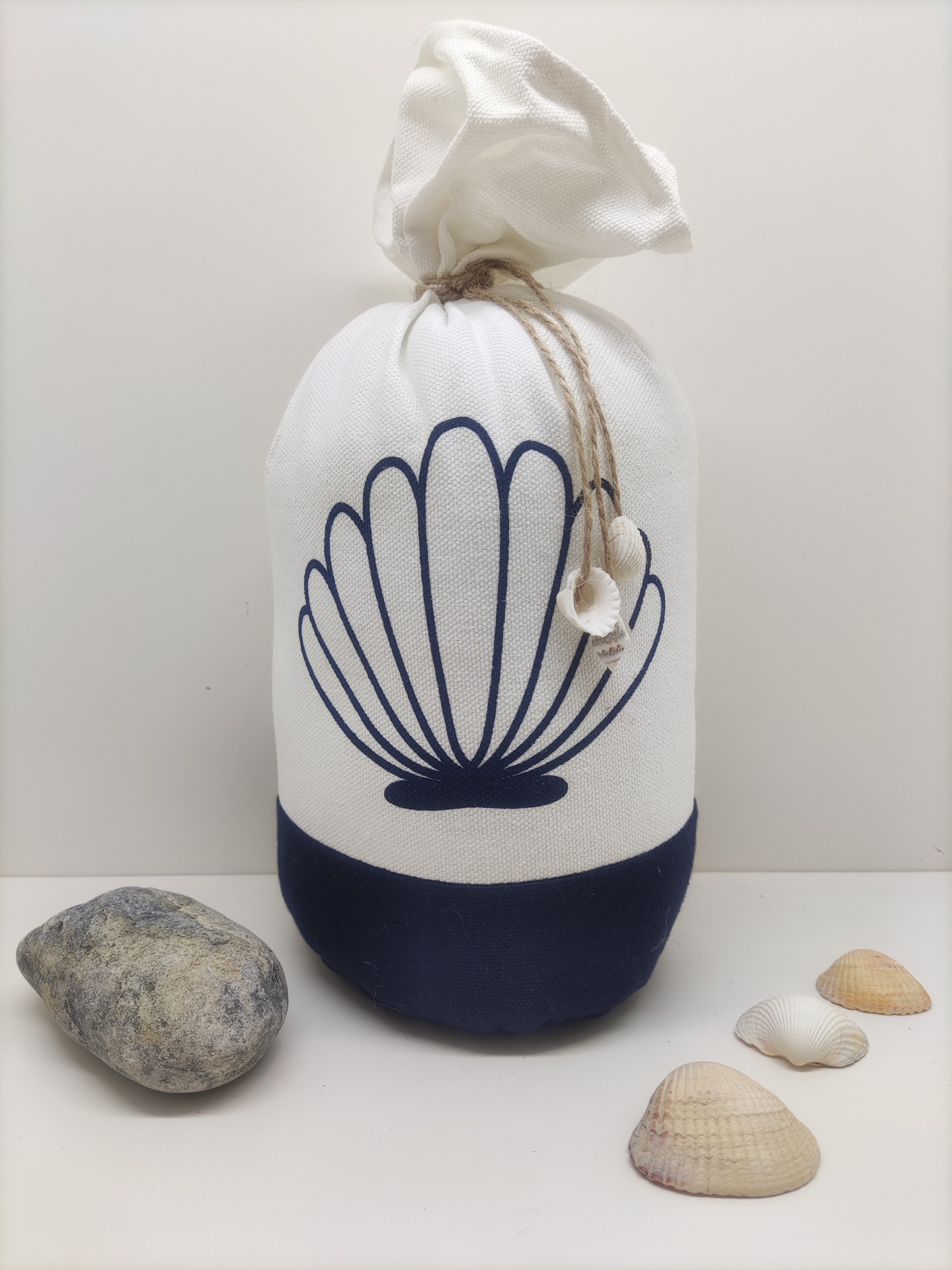 Saco tecido branco e azul com desenho de concha e cordão, conchas e pedra ao redor.