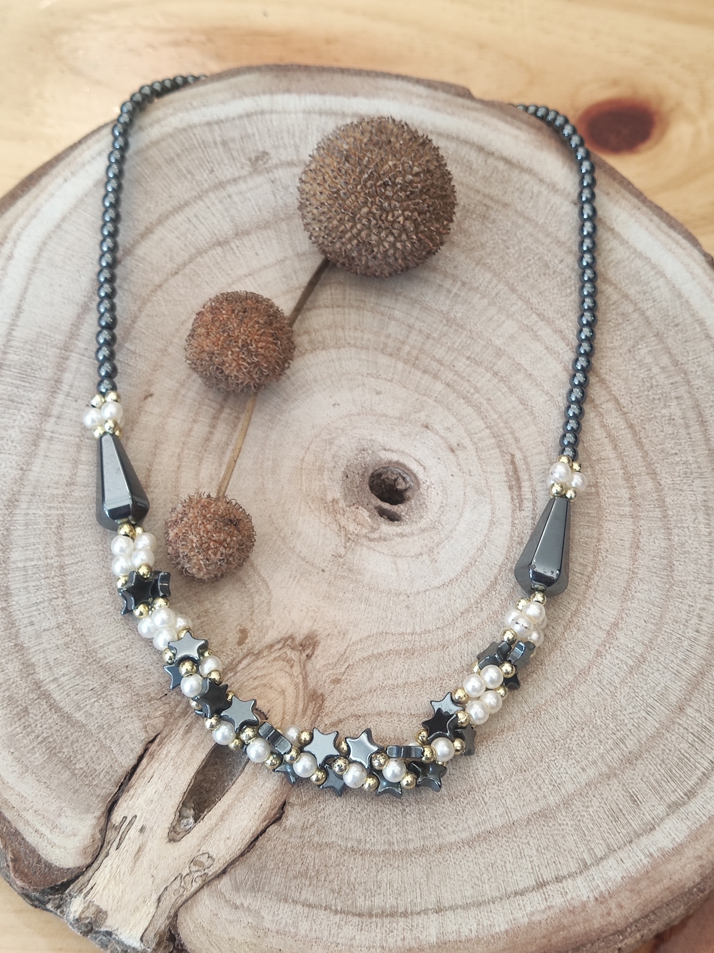 Colar com contas pretas, pérolas brancas e estrelas metálicas sobre base de madeira redonda com sementes secas