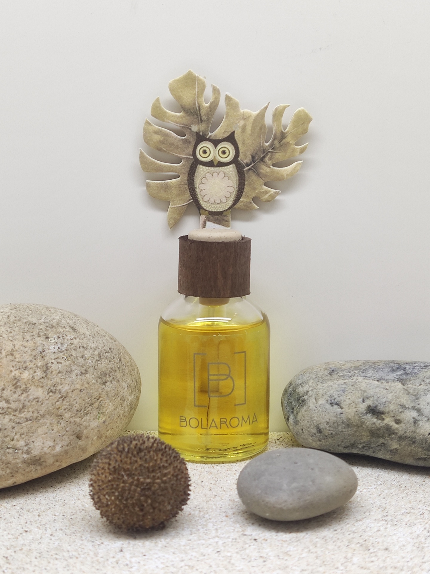 Frasco de difusor de aroma com tampa decorada por coruja, rodeado por pedras sobre superfície clara