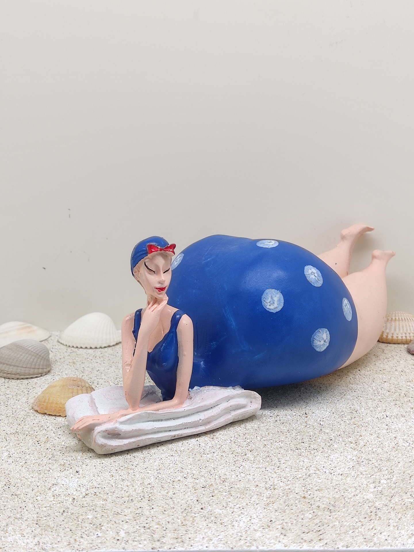 Escultura de mulher de fato de banho azul com bolas brancas, deitada na toalha, com conchas ao redor.