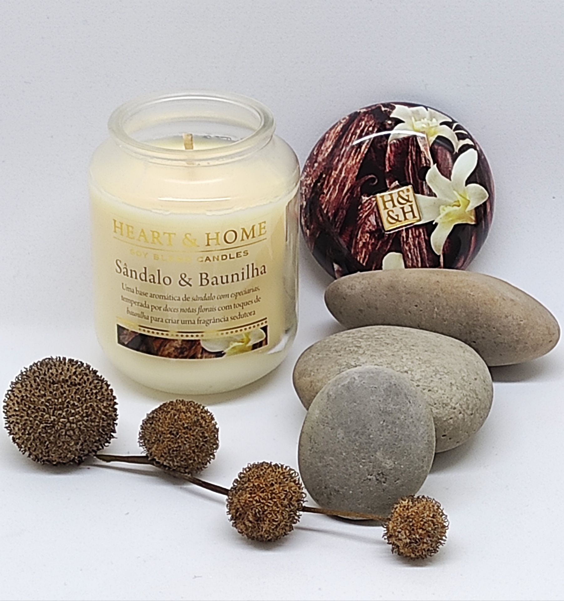 Vela perfumada em frasco de vidro com tampa decorativa ao lado de pedras lisas e frutos secos