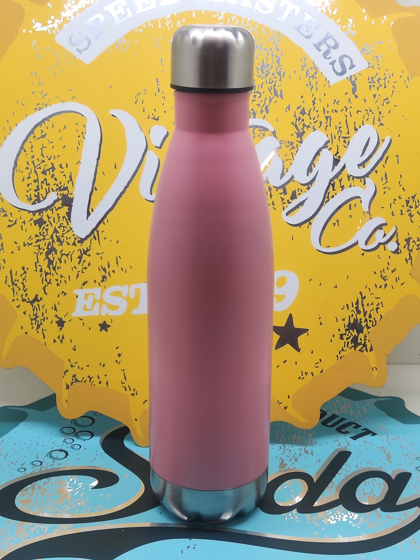 Garrafa térmica rosa com tampa prateada sobre fundo amarelo com texto e superfície azul decorada