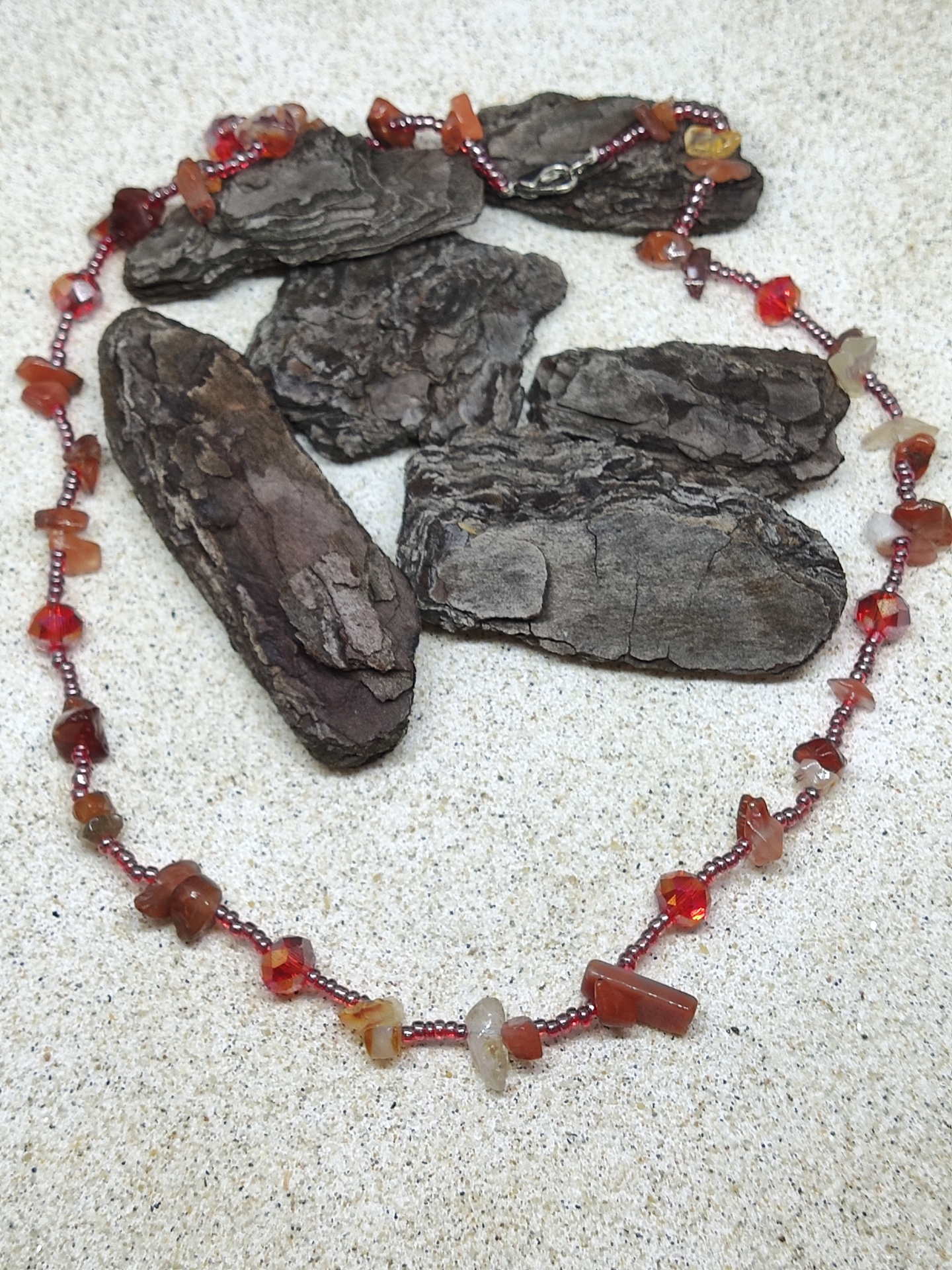 Colar de pedras naturais vermelhas, castanhas e contas vermelhas em superfície bege com casca de árvore.