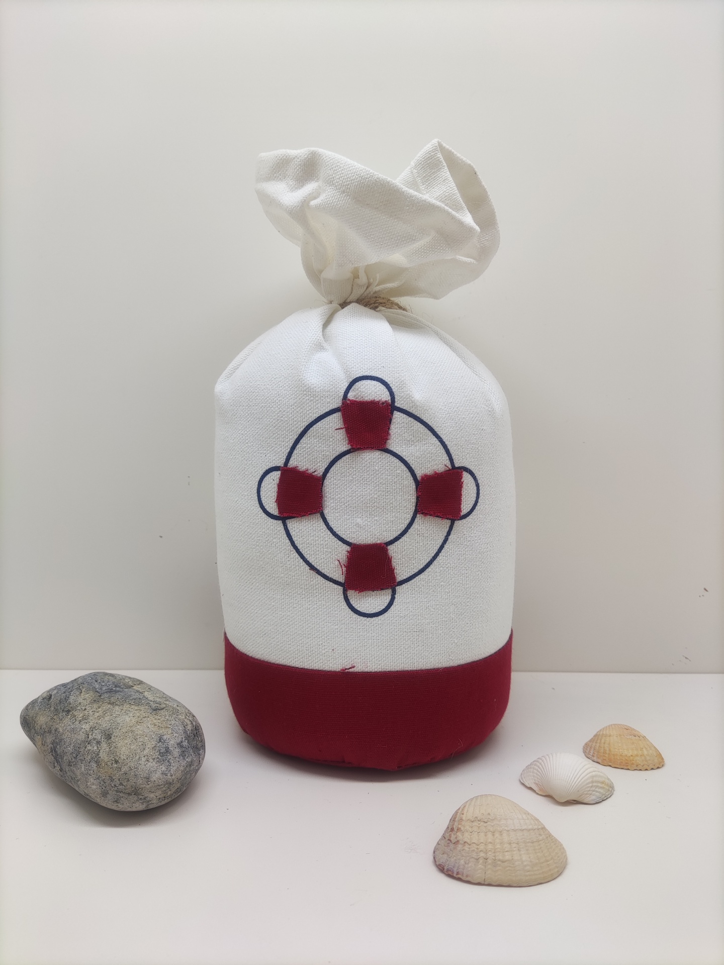 Saco branco e vermelho com desenho de bóia salva-vidas, conchas e pedra ao lado.