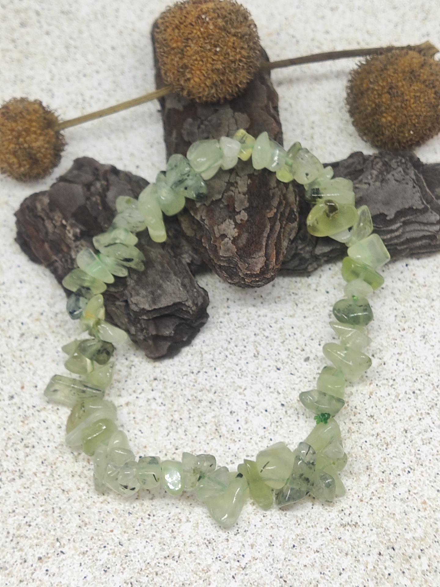 Pulseira de pedras verdes translúcidas com textura irregular em fundo cinza com casca de árvore e flores secas