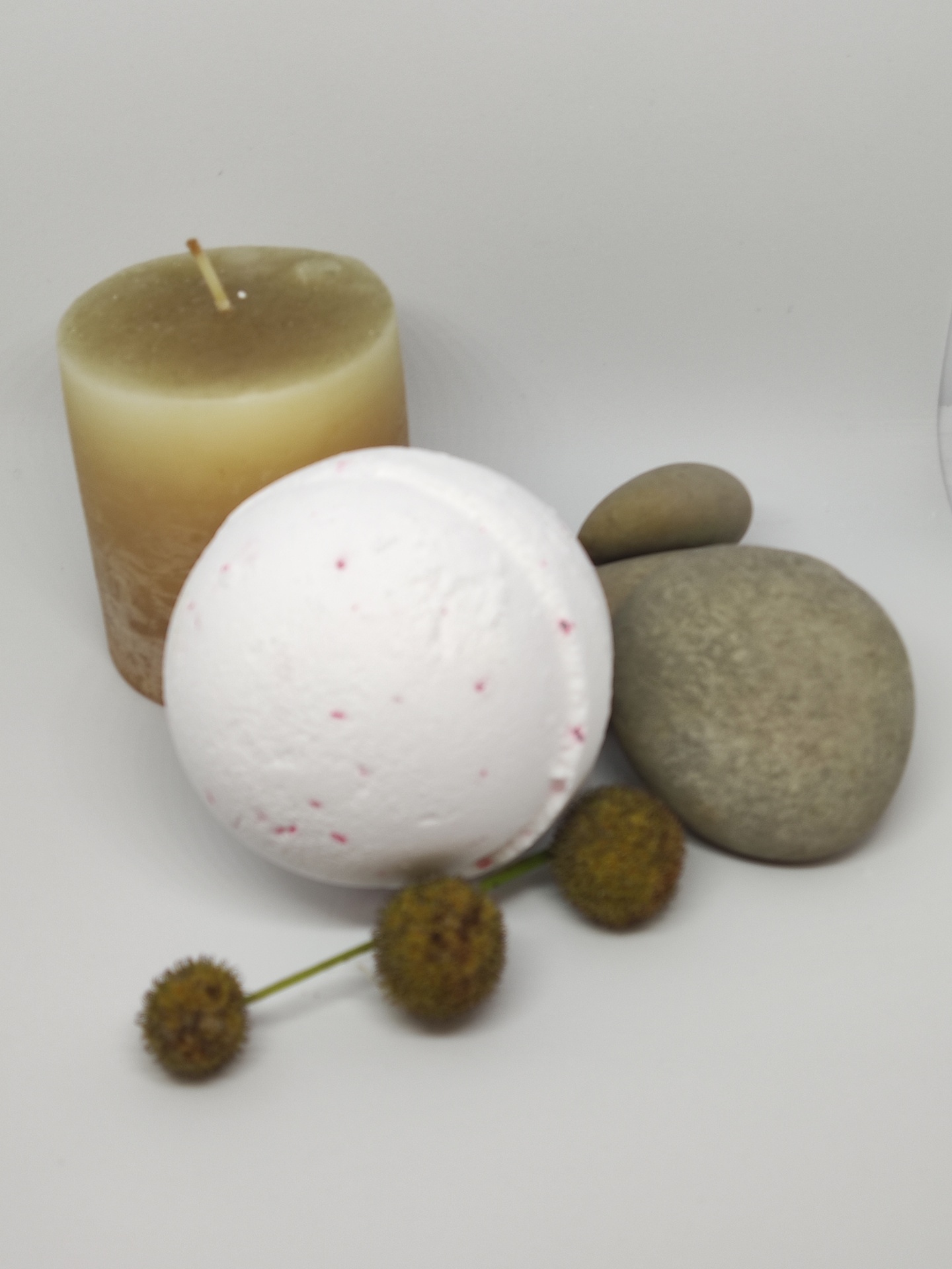 Vela creme, bomba de banho branca com pontos vermelhos, pedras cinzentas e flores secas sobre superfície branca