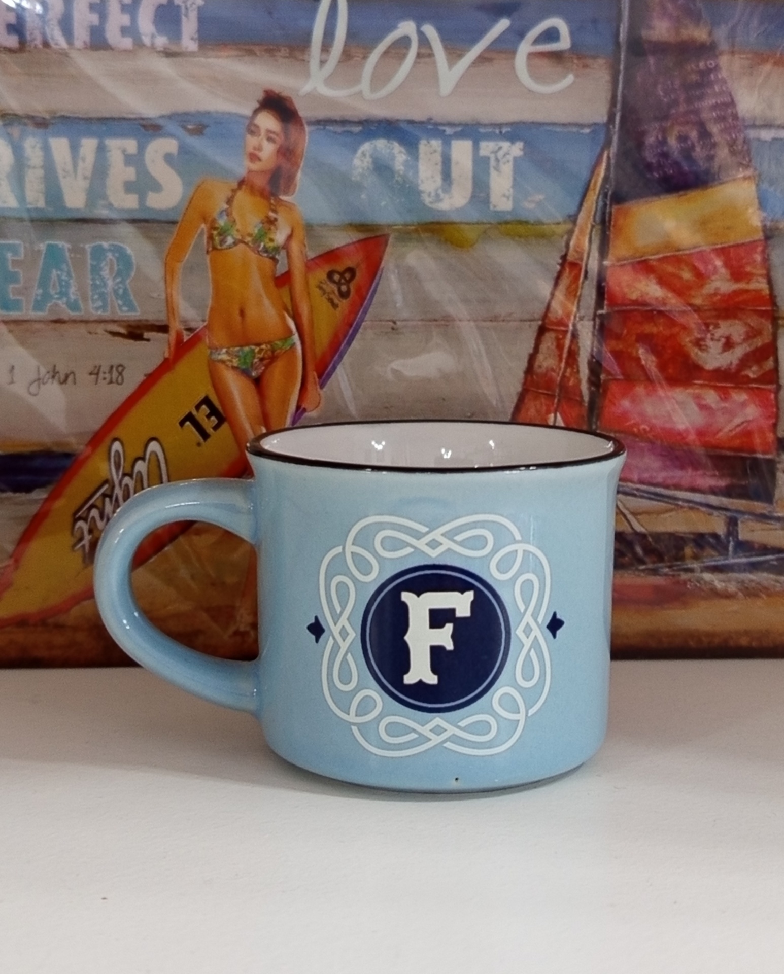 Caneca azul claro com letra F em círculo azul e padrão decorativo, em superfície branca e fundo com imagem colorida de mulher com prancha de surf.