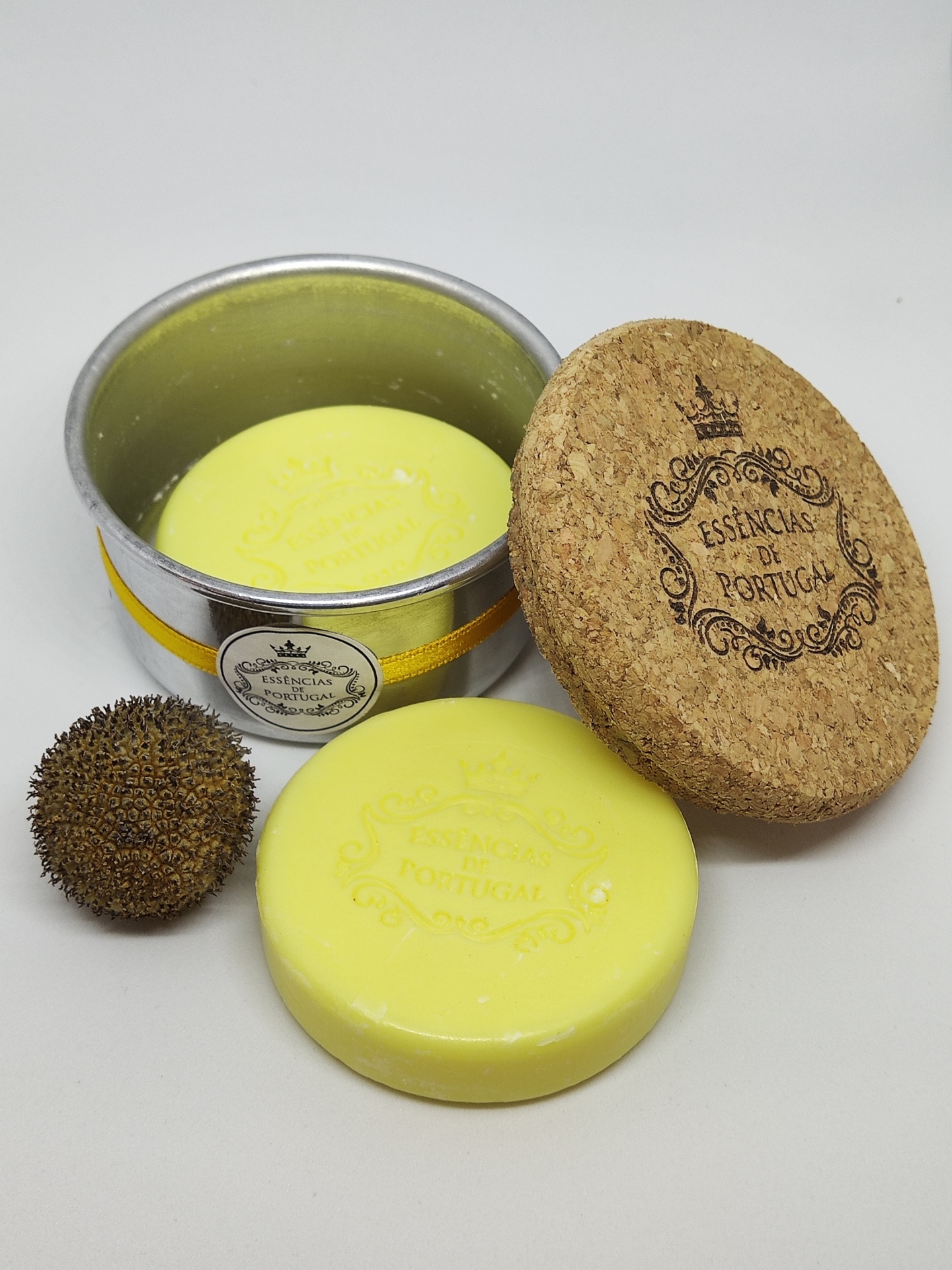 Sabonete amarelo redondo com embalagem metálica e tampa de cortiça, com esfera marrom texturada