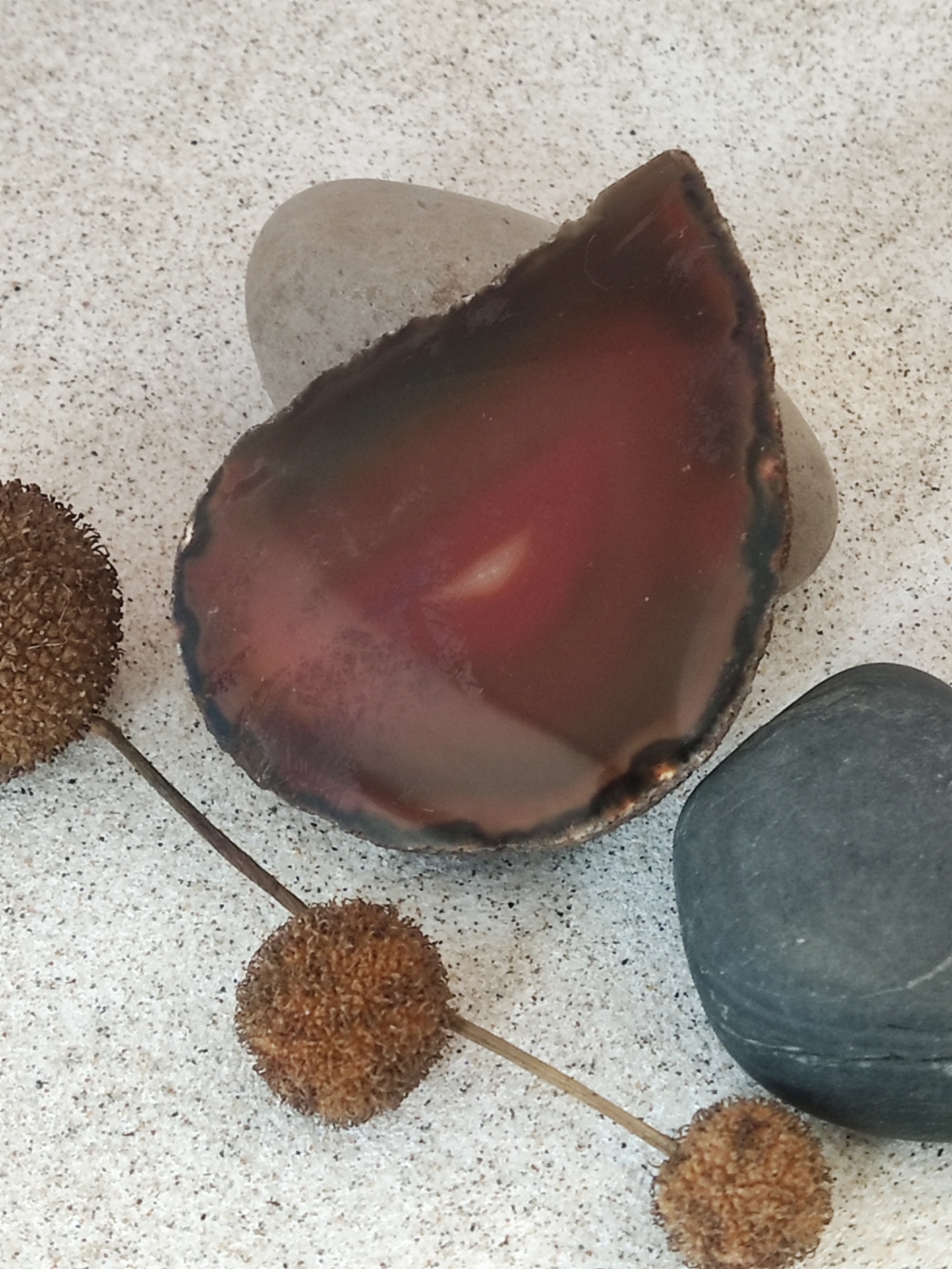 Ágata polida castanha e preta sobre pedras cinzentas com sementes secas marrons num fundo claro.