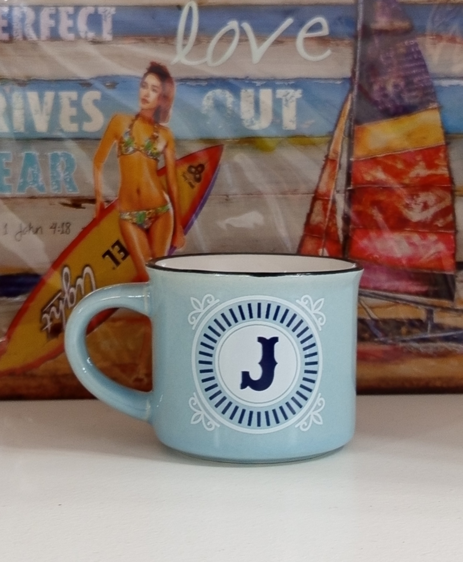 Caneca azul clara com a letra J em círculo, frente a fundo com mulher de biquíni e prancha de surf.