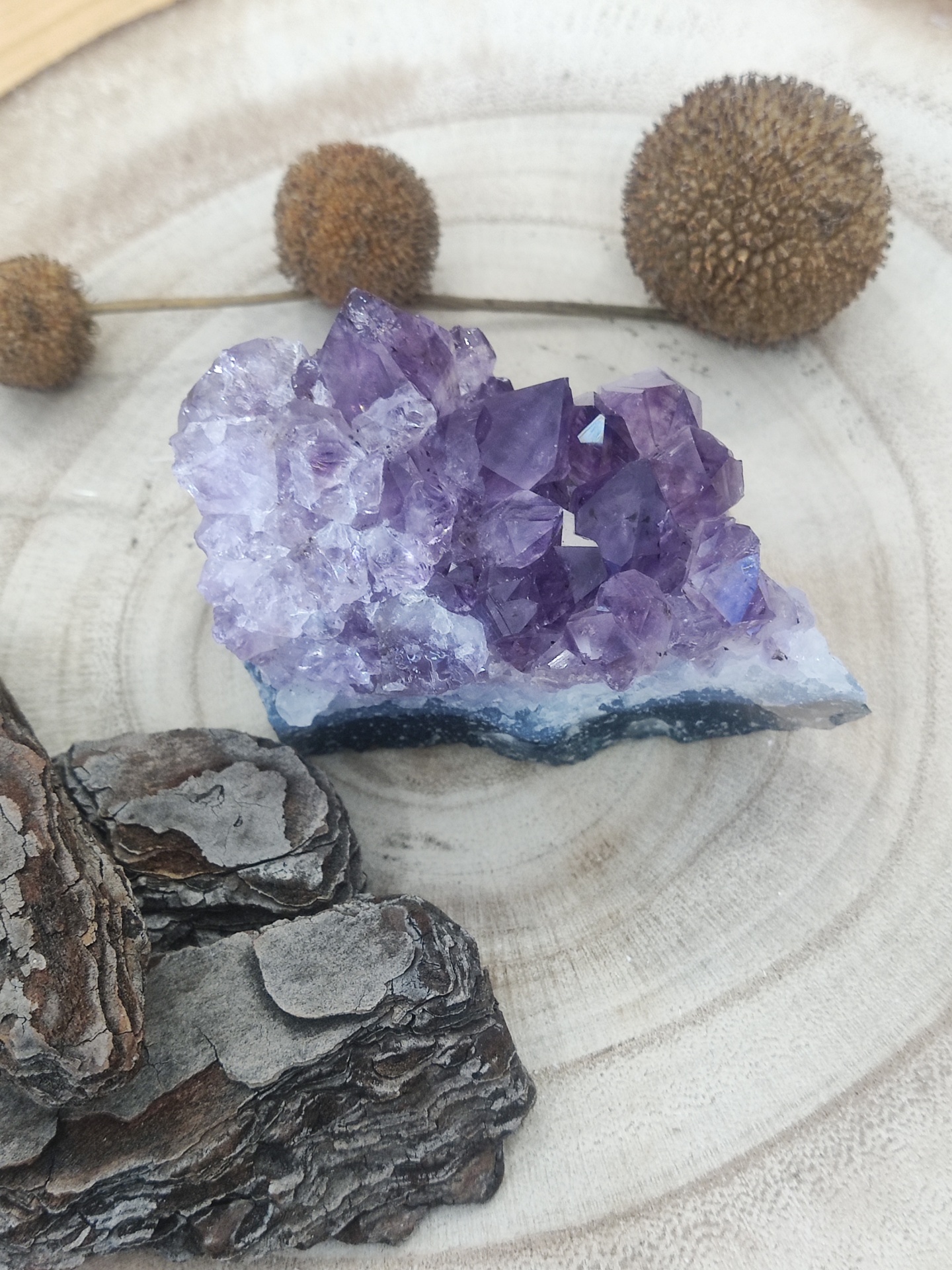 Cristal de ametista roxo sobre madeira clara com casca de árvore e sementes secas arredondadas.