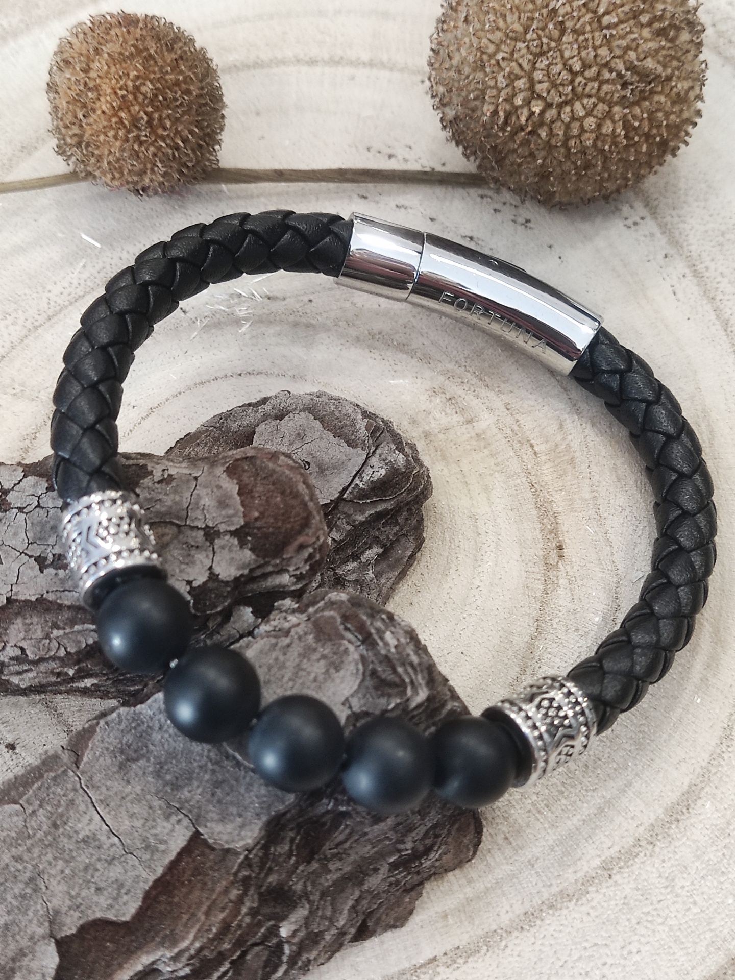 Pulseira preta em couro trançado com contas negras e detalhes prateados sobre casca e fundo claro