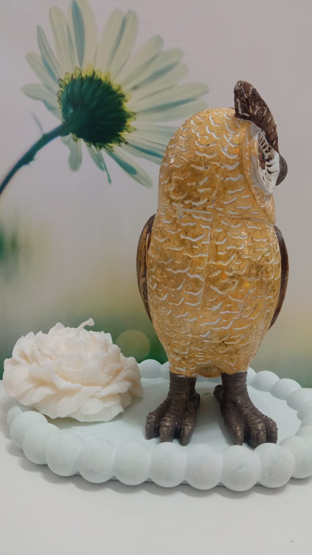 Estatueta de coruja dourada e castanha numa base branca com flor branca e fundo floral