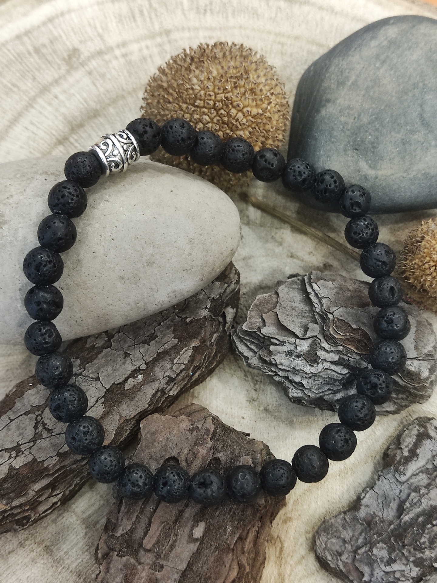 Pulseira de contas negras de lava vulcânica com detalhe metálico prateado sobre pedras e casca seca