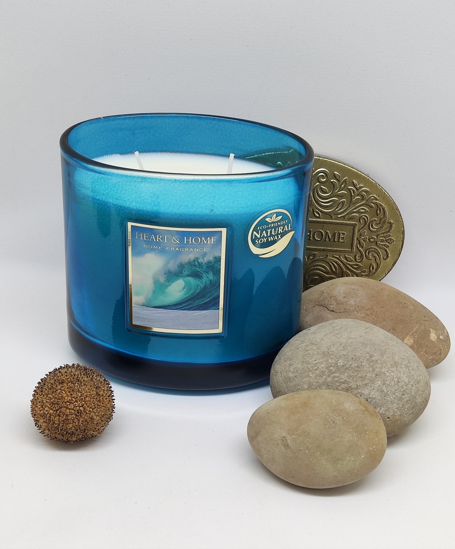Vela perfumada em vidro azul com três pavios e etiqueta com onda junto a pedras lisas e bola castanha