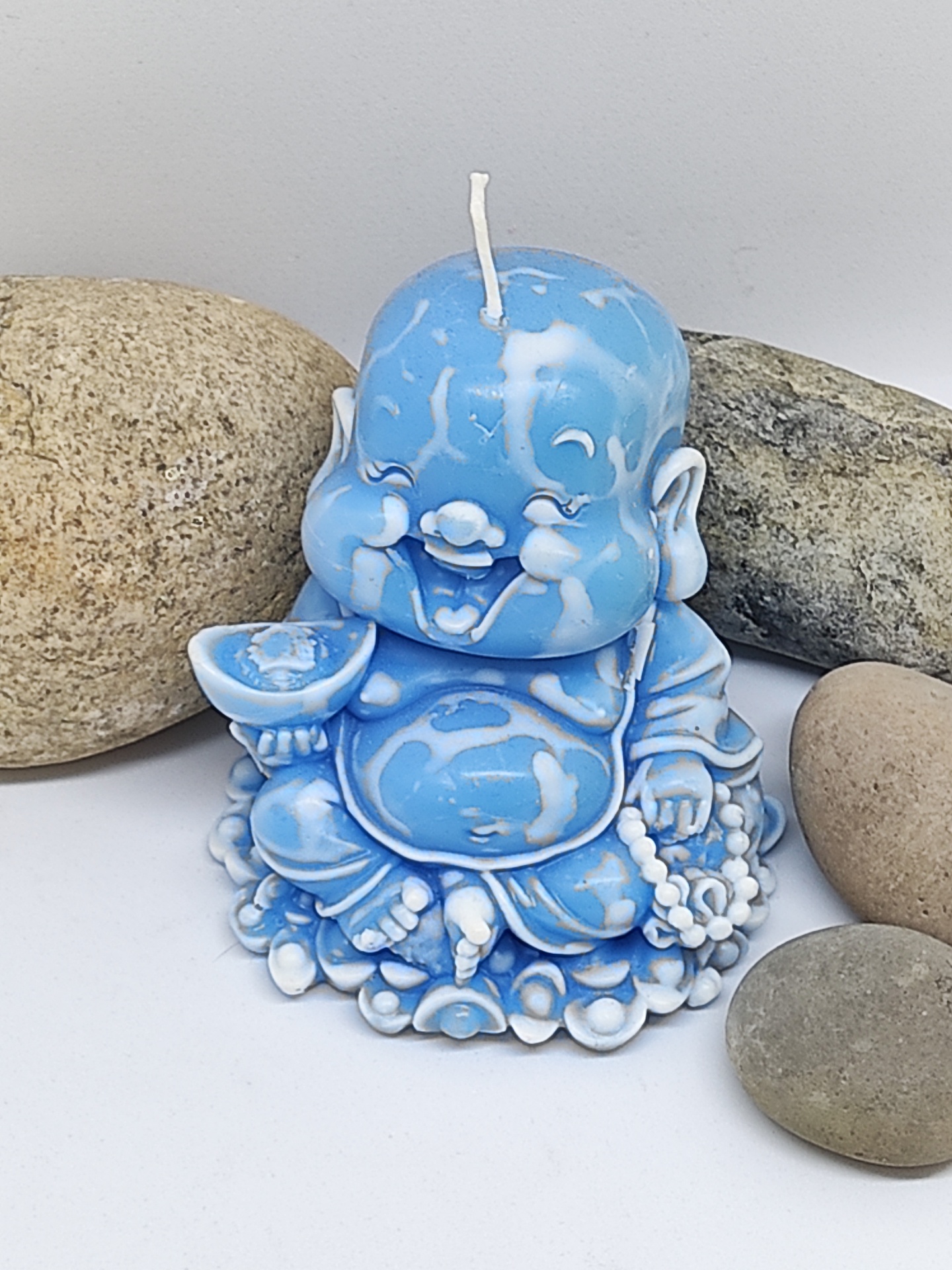 Vela azul e branca em forma de Buda sorridente rodeada por pedras cinzentas.