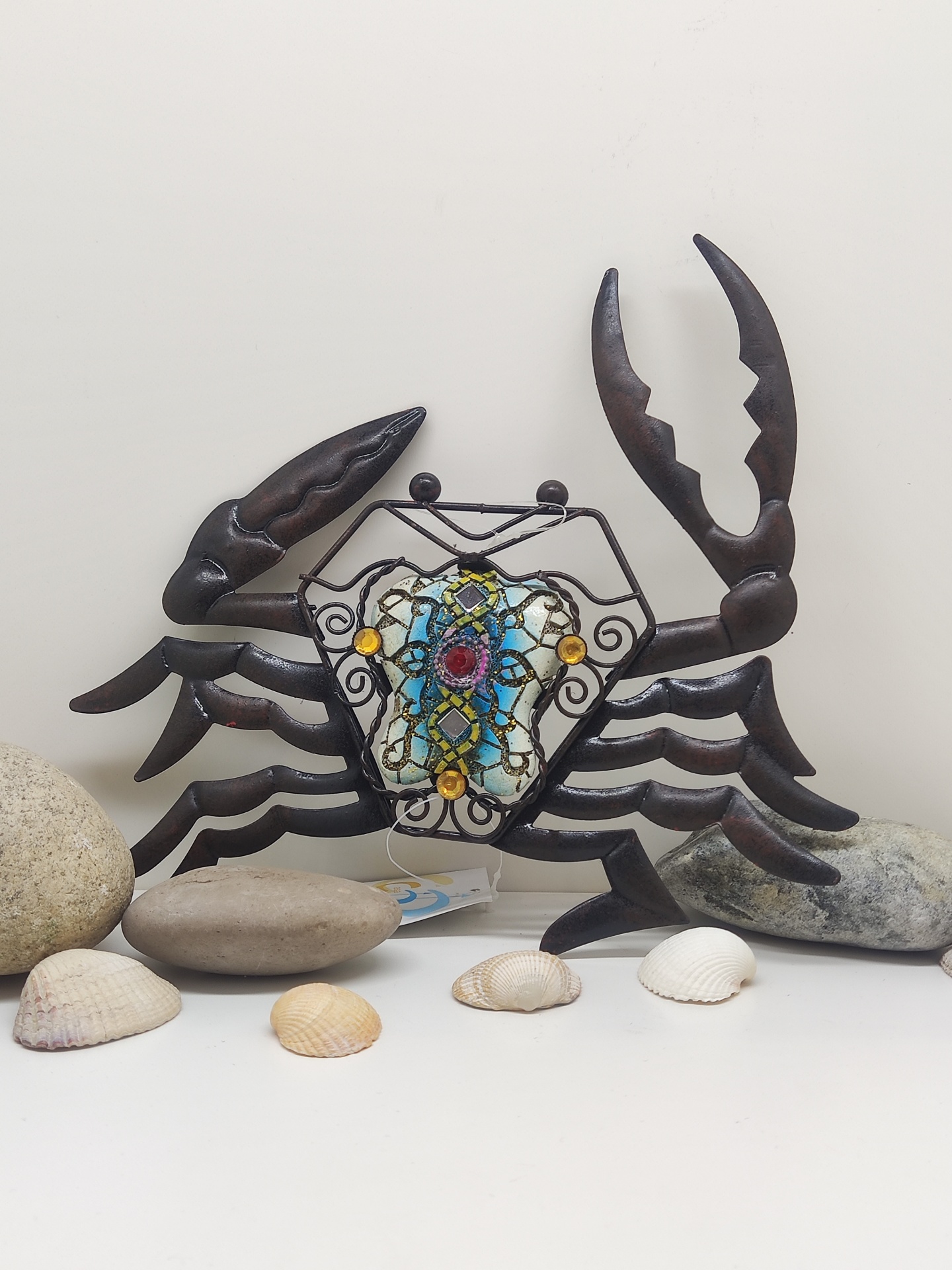 Escultura decorativa de caranguejo em metal com vidro colorido rodeada por conchas e pedras