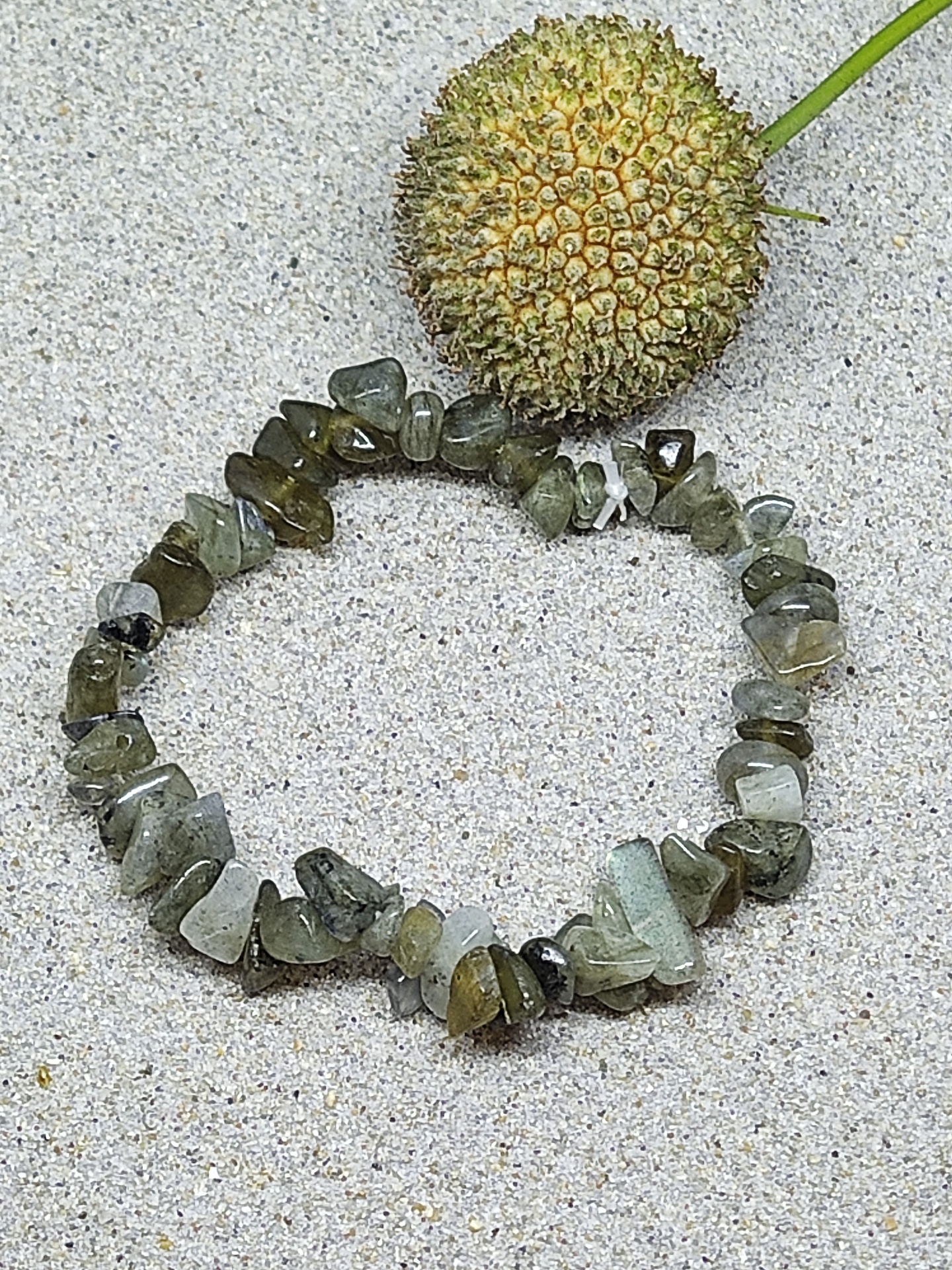 Pulseira verde de pedras irregulares sobre areia ao lado de fruto seco espigado verde