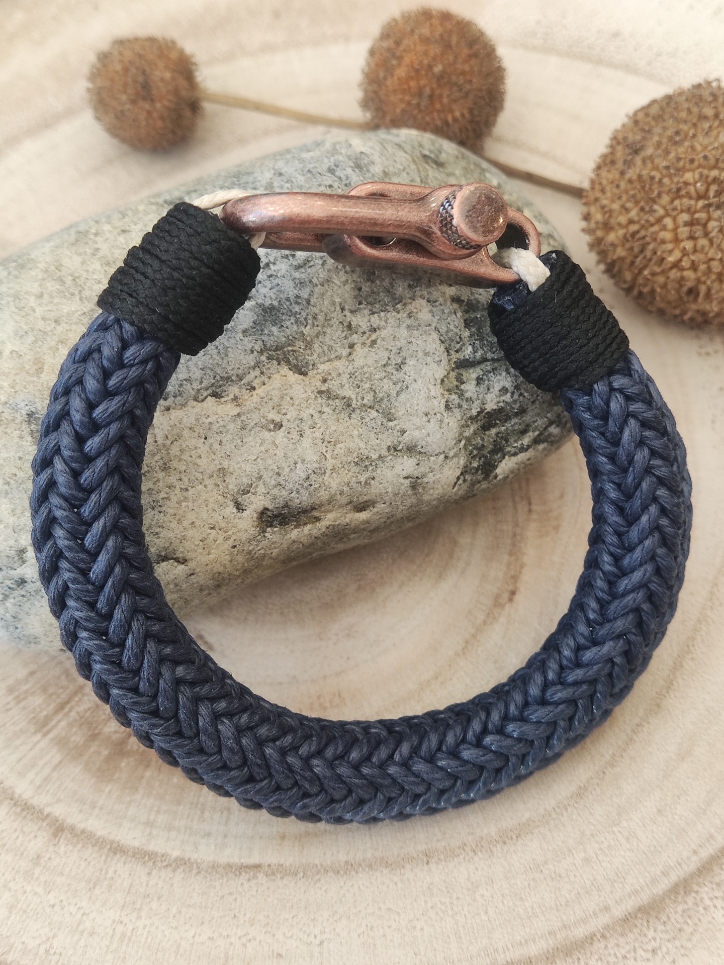 Pulseira de corda azul marinho trançada com fecho metálico cor de cobre sobre pedra cinza e fundo de madeira.