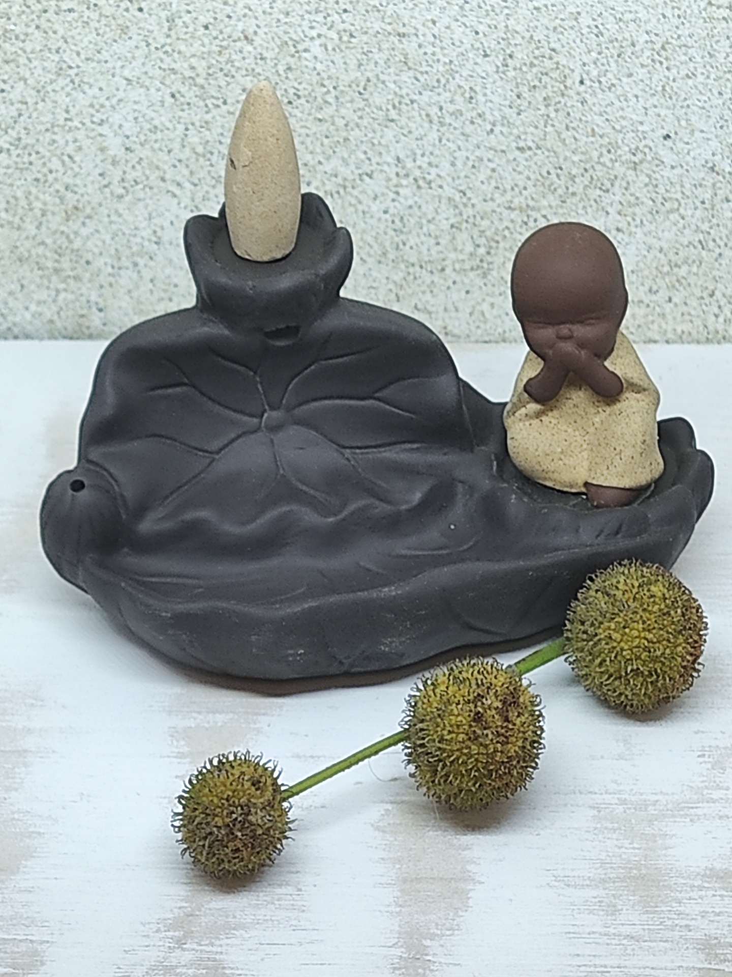 Incensário cerâmico em forma de folha com monge e cone de incenso, decorado com flores esféricas.