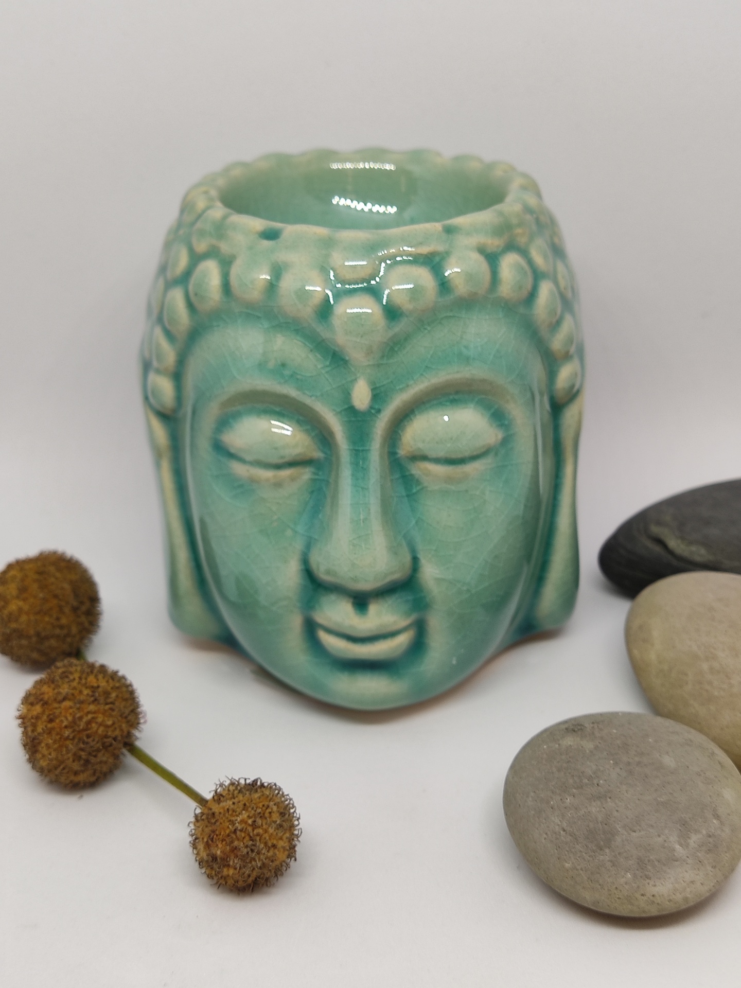Vela aromática em cerâmica azul esverdeada com formato de cabeça de Buda rodeada por pedras e flores secas