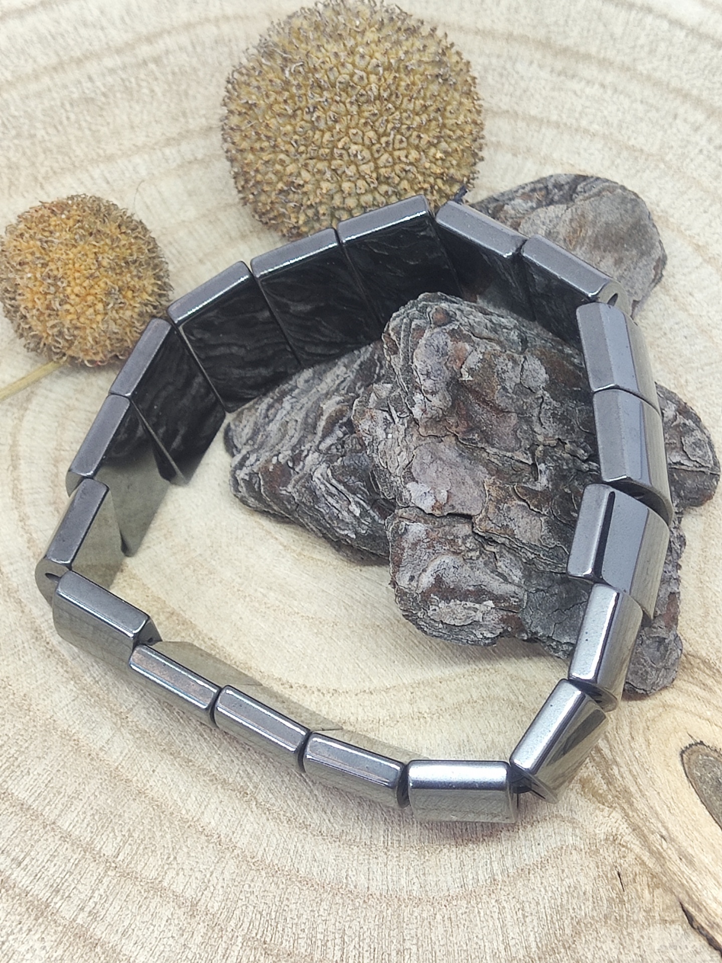 Pulseira metálica cinza escuro com peças retangulares sobre madeira e sementes secas