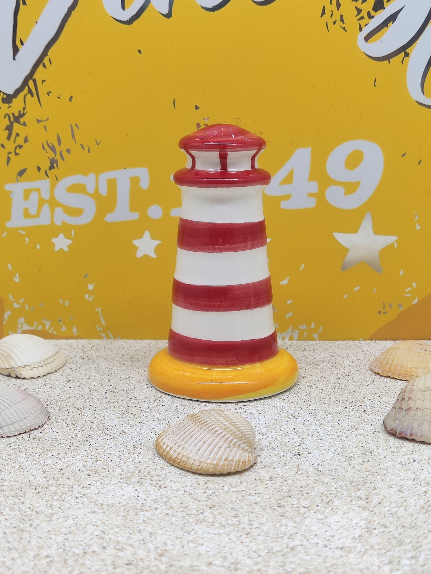 Miniatura decorativa de farol listrado vermelho e branco com base amarela e conchas ao redor