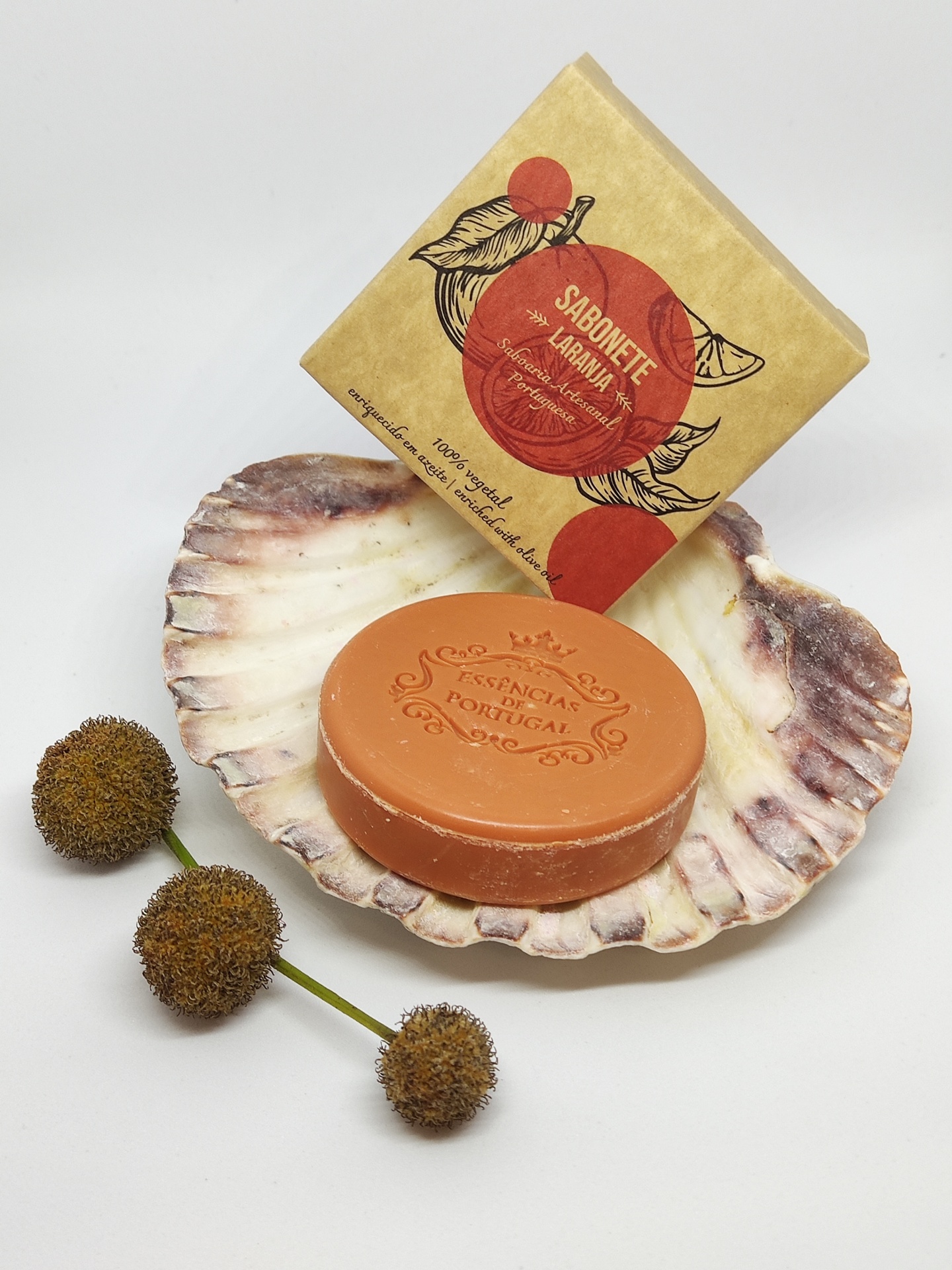 Sabonete laranja redondo numa concha com embalagem de papel e flores secas ao lado