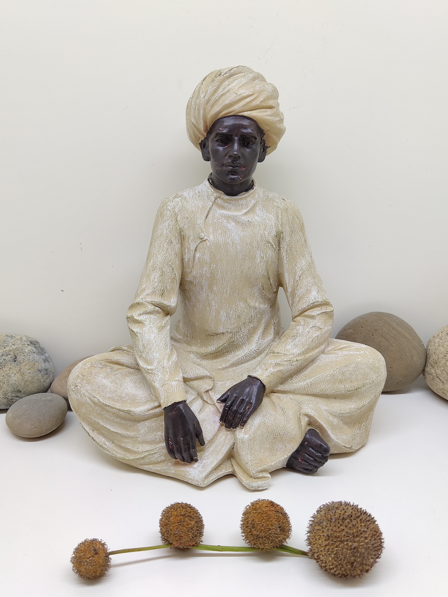 Estátua decorativa de homem sentado com turbante e roupa bege, com pedras e frutos secos ao redor.