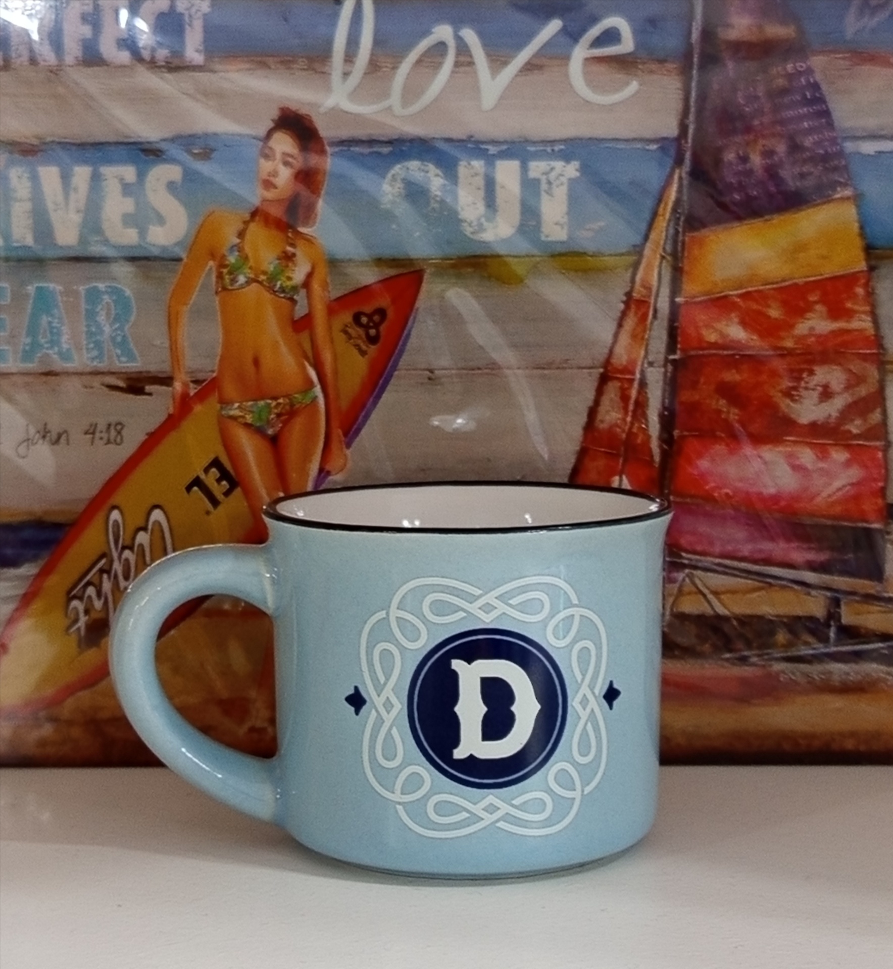 Caneca azul claro com letra D em fundo com pintura de mulher de biquíni e prancha de surf