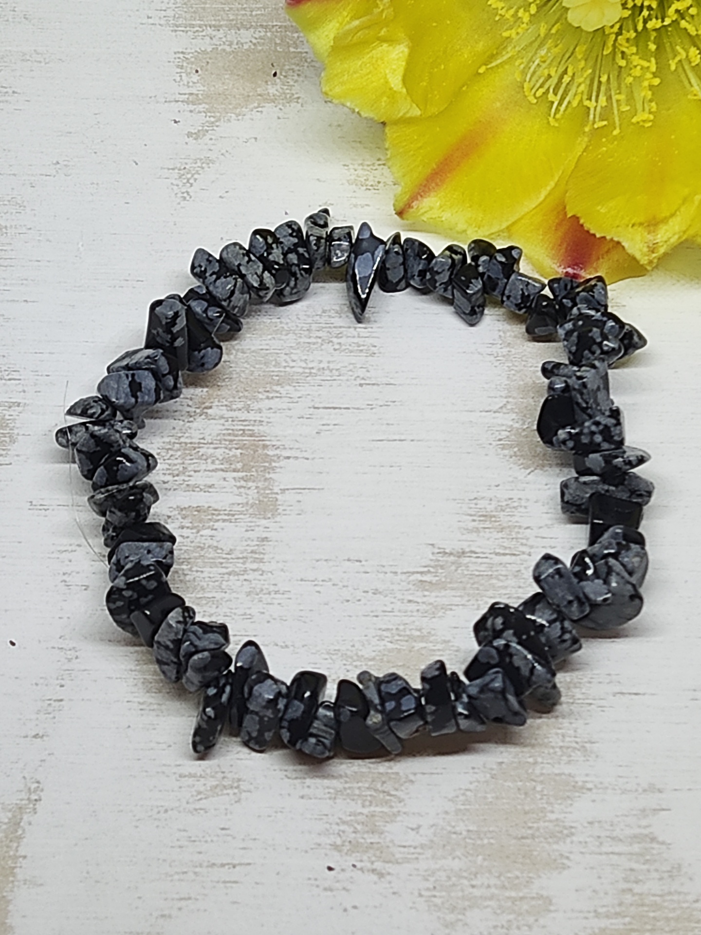 Pulseira de pedras pretas e cinzentas com padrão irregular sobre fundo de madeira clara e flores amarelas