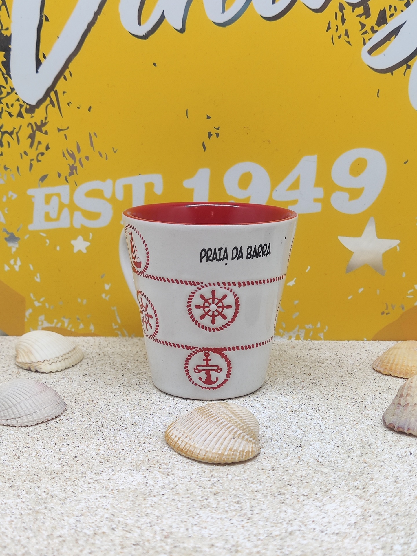 Caneca cerâmica vermelho e branco com motivos náuticos e texto Praia da Barra sobre areia com conchas e fundo amarelo com texto.