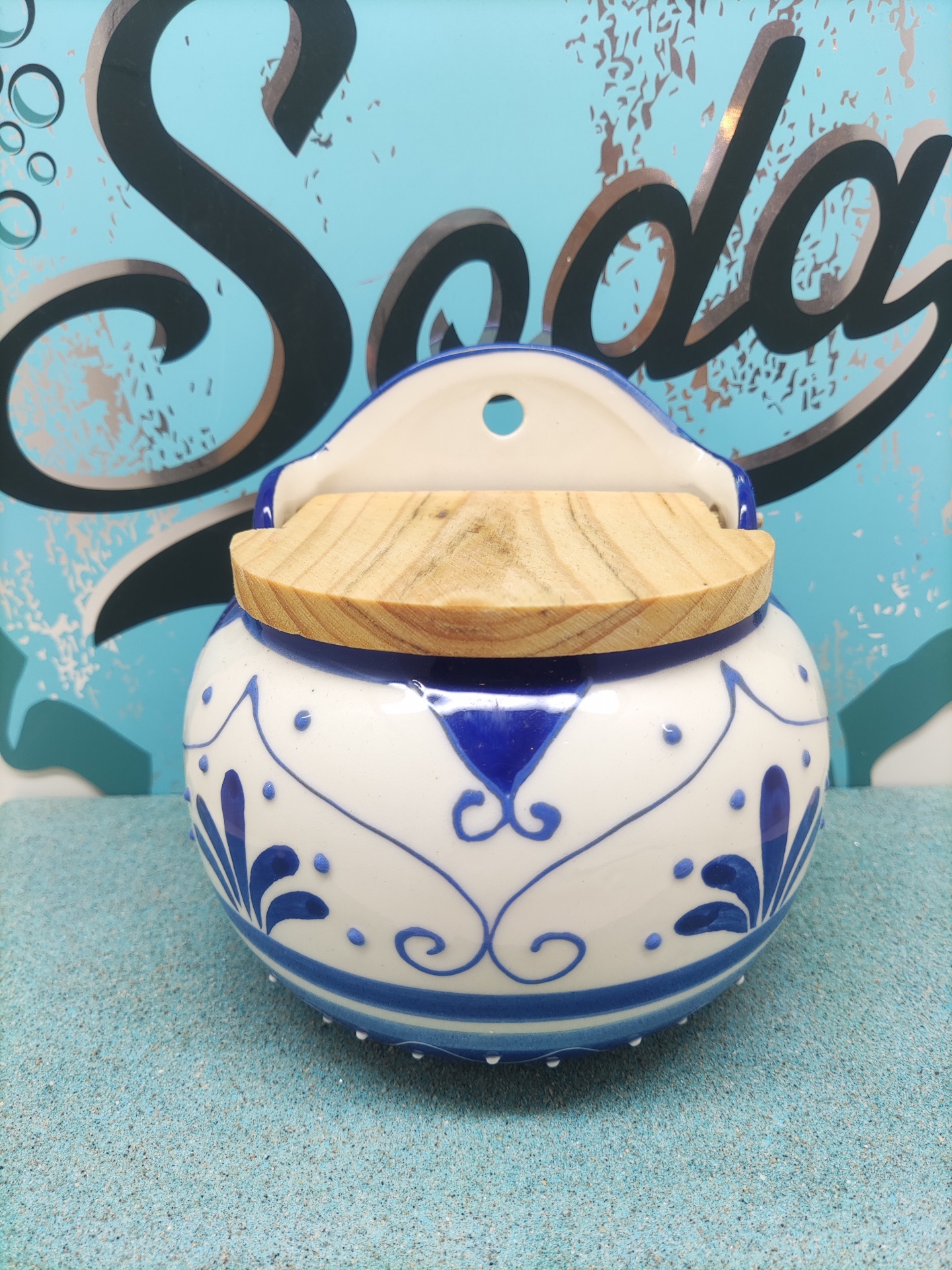 Recipiente de cerâmica branca e azul com tampa de madeira e fundo com texto Soda